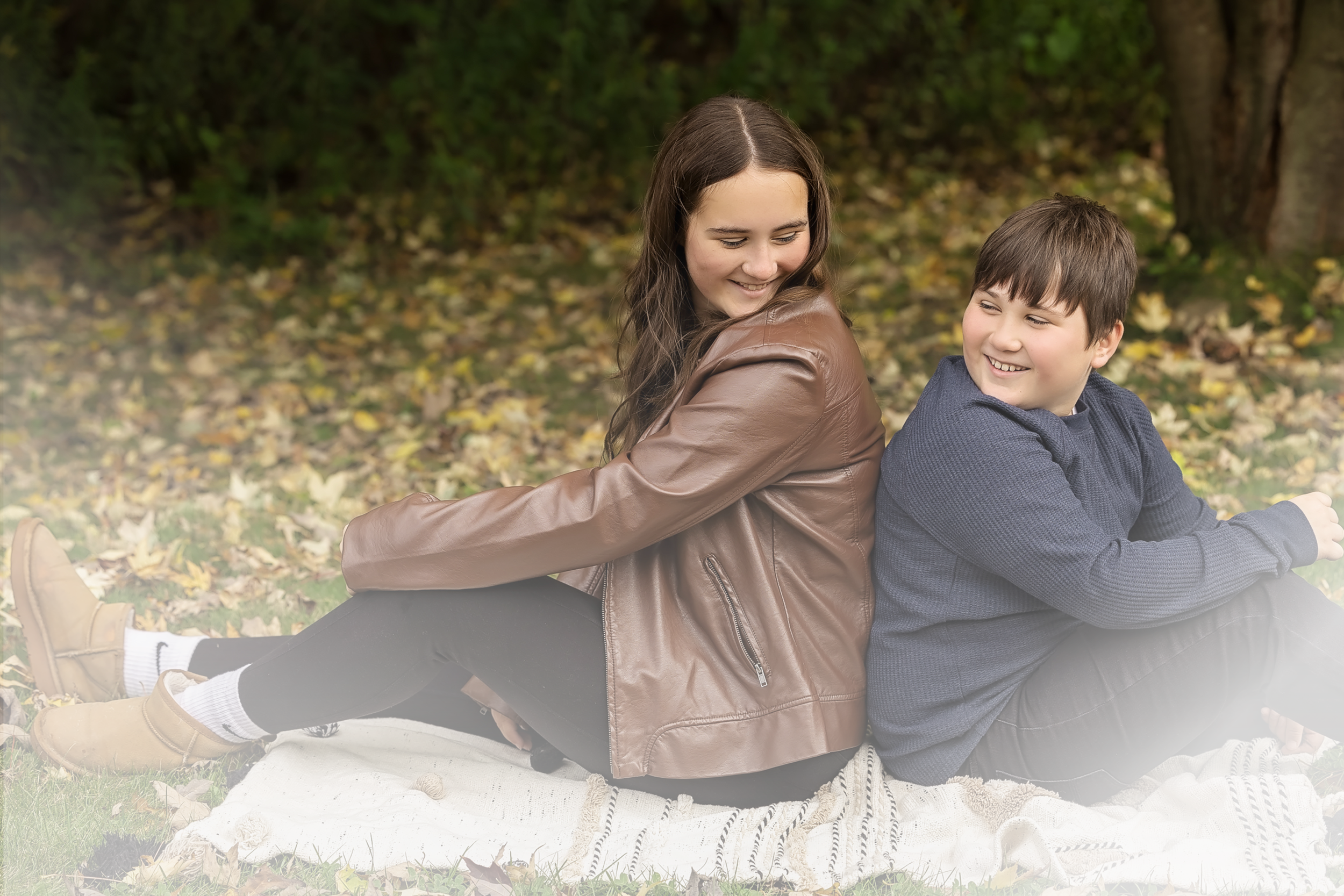 A smiling teenage girl with long brown hair and a brown leather jacket sitting back-to-back with a young boy with short brown hair and a blue knitted sweater outdoors in a park with autumn leaves on the ground and trees in the background.