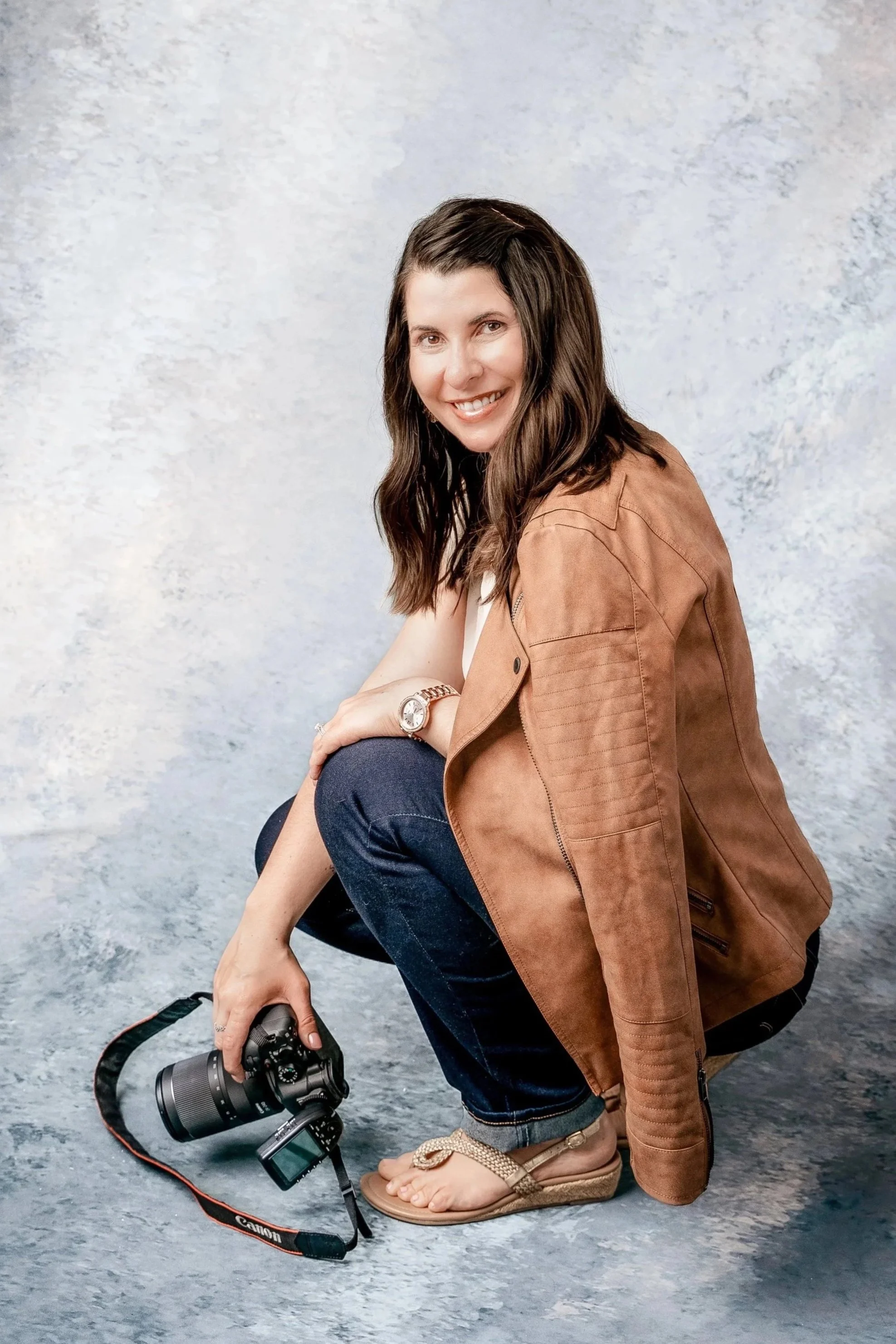 A woman with long brown hair, wearing a brown jacket and dark blue jeans, is crouching down while holding a camera. She is smiling and looking at the camera, with a light gray textured background behind her.