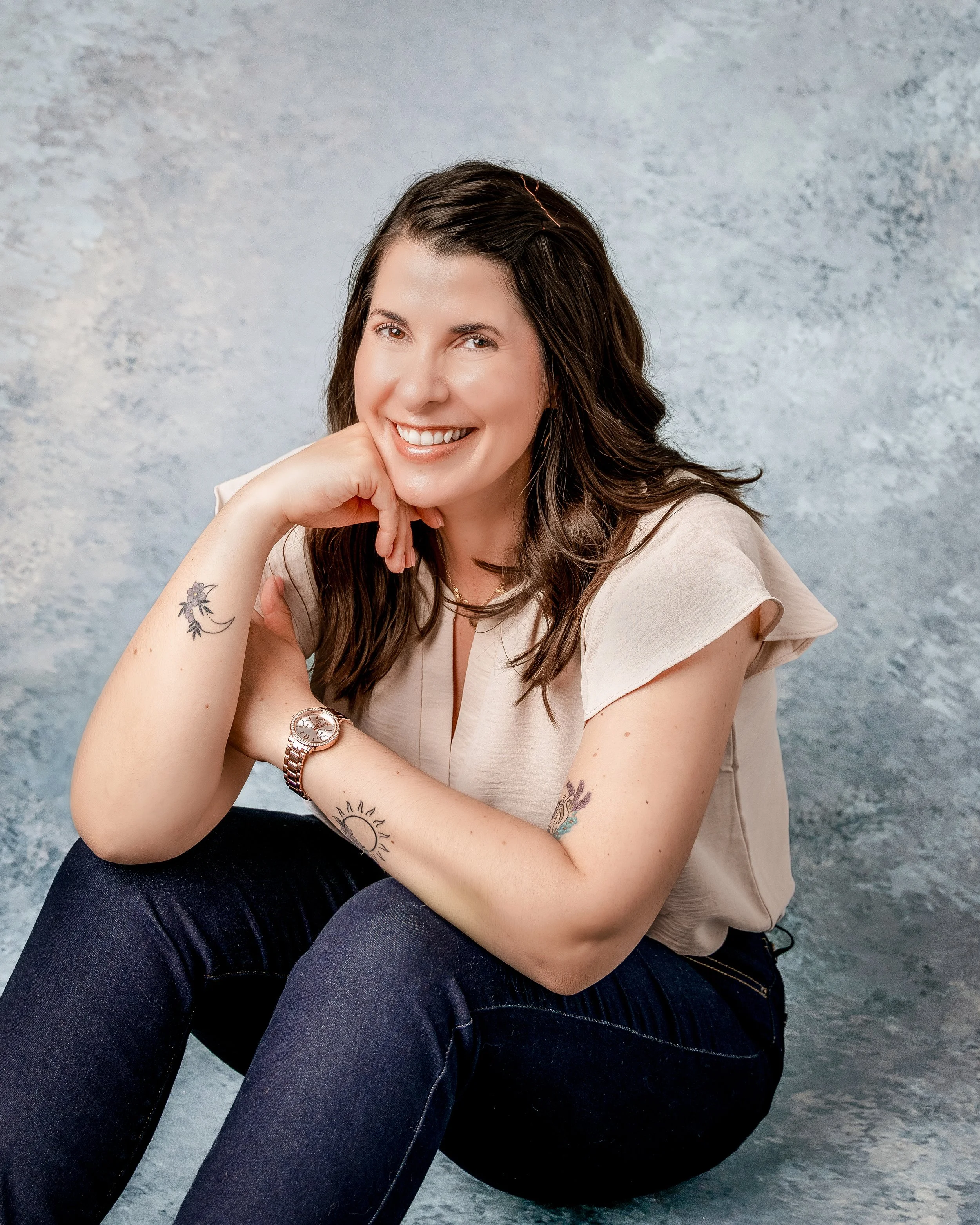 A woman with long brown hair and tattoos smiling and resting her chin on her hand, wearing a beige top, dark jeans, gold jewelry, on a textured gray background.
