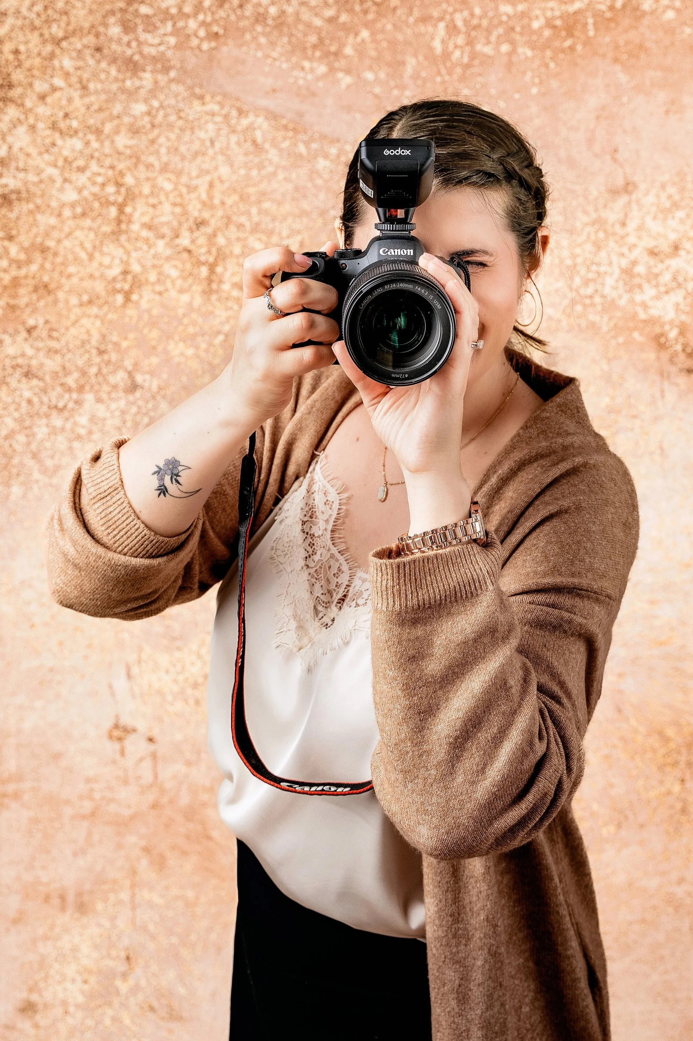 A woman taking a photo with a Canon camera, wearing a beige cardigan, white blouse, and black pants, standing against a textured beige wall.