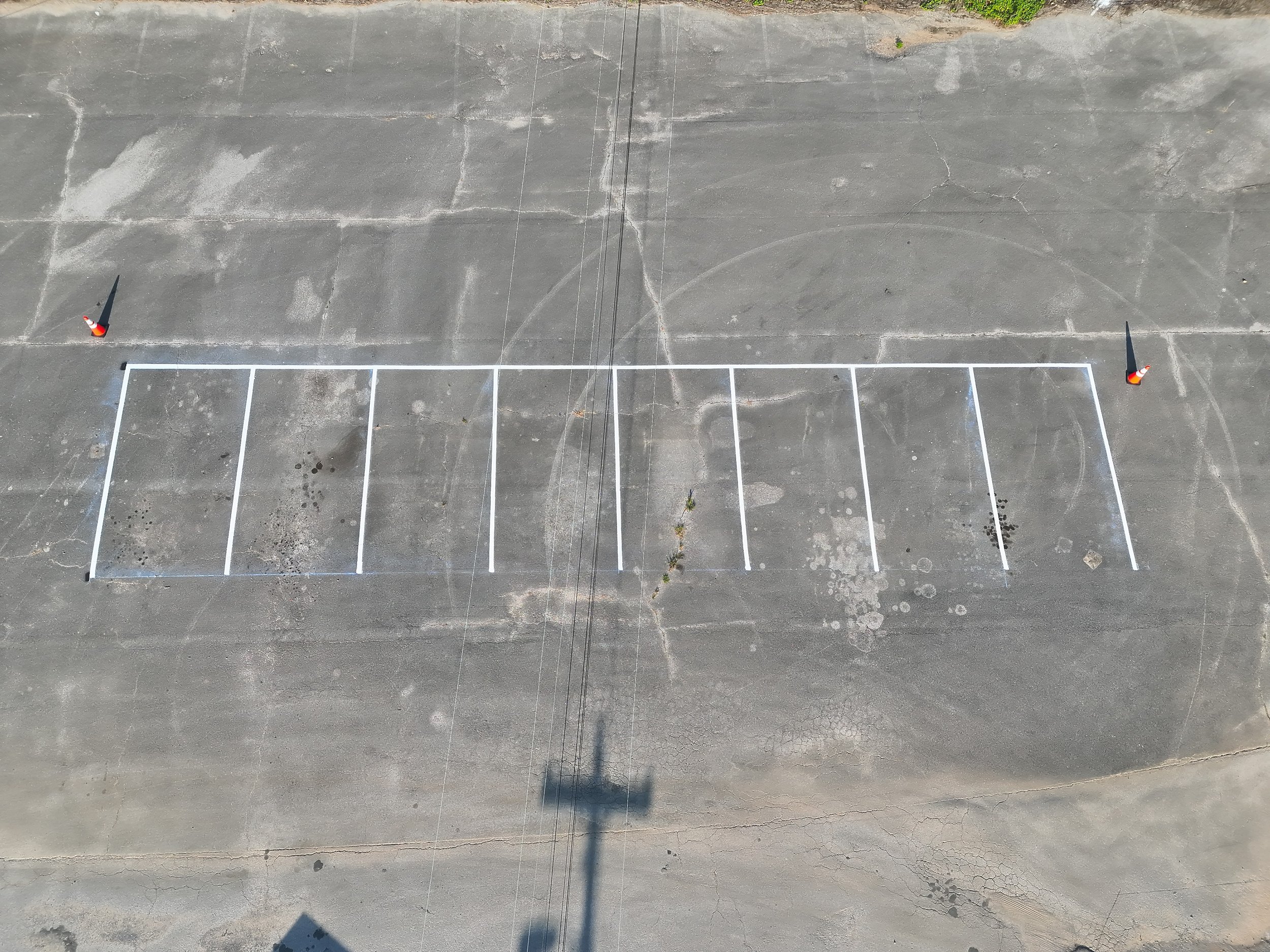 An aerial view of an outdoor basketball court with white lines; orange traffic cones are placed at the two ends of the court.