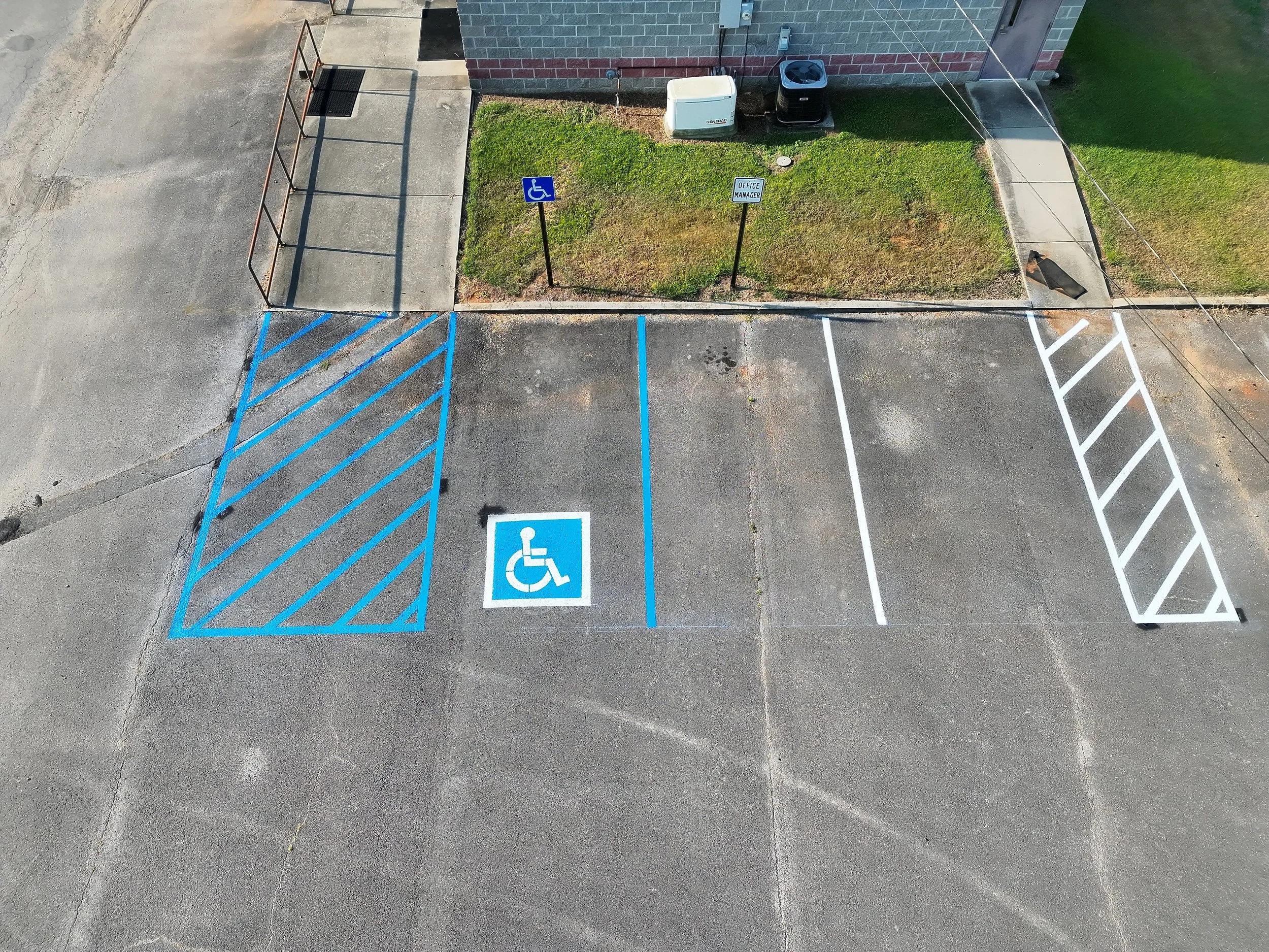 An aerial view of a parking lot showing a designated accessible parking space with blue and white markings and a handicapped symbol, next to a small grass area with two signs that say 'Accessible Parking' and 'Office Manager.' There are concrete sidewalks, a brick building, and some utilities nearby.