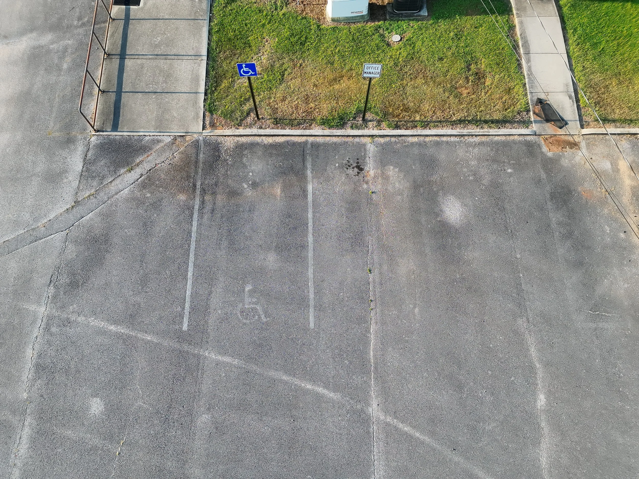 An aerial view of a parking lot with a small grassy area in the center. The grassy area has two signs: a handicap parking sign and an office manager sign. The parking lot has designated space lines and some cracked asphalt. There are also some utility boxes and electrical wires visible.