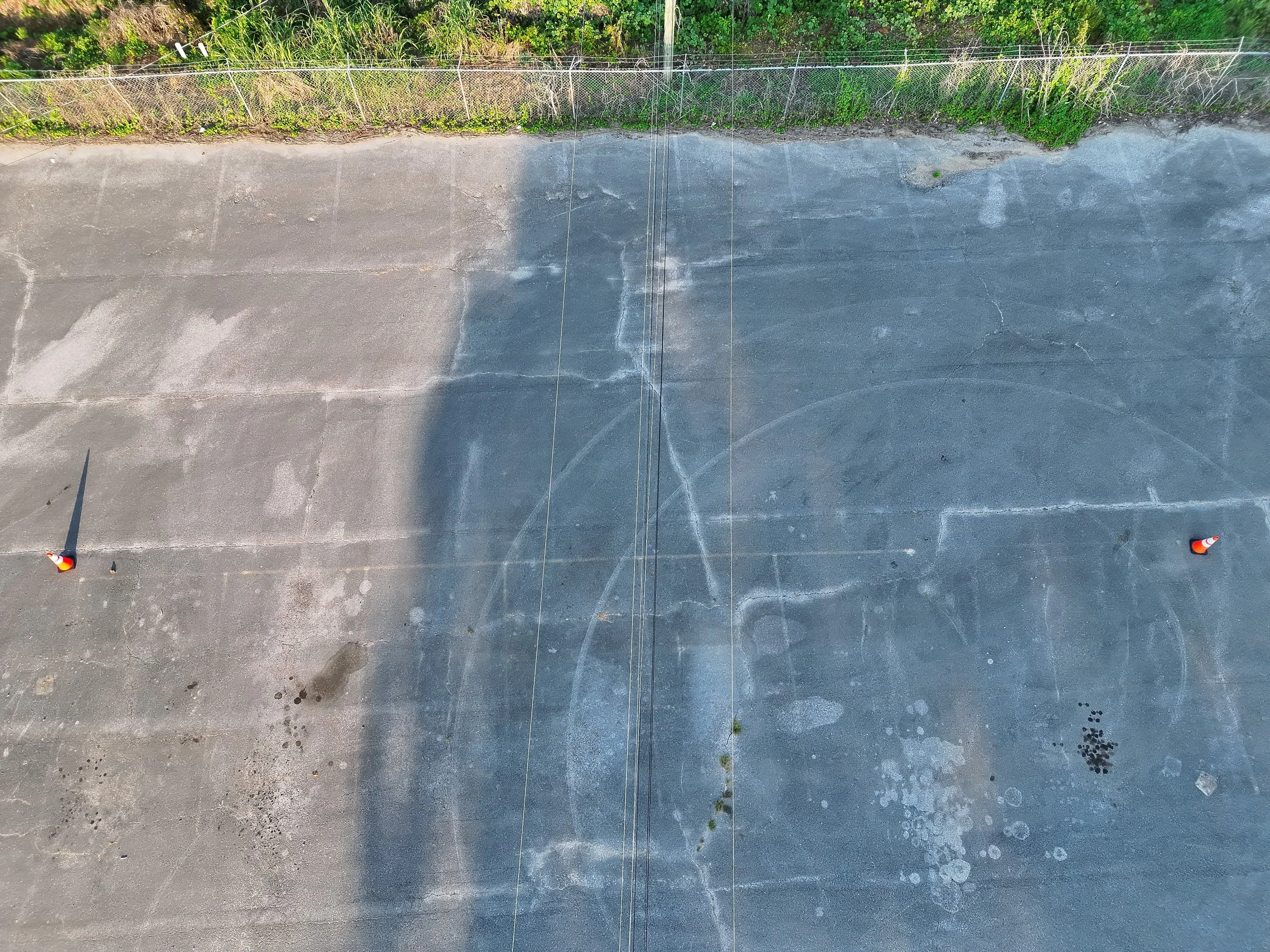 Aerial view of a parking lot with faint markings, some wet spots, and orange traffic cones at the bottom corners, bordered by grass and plants.