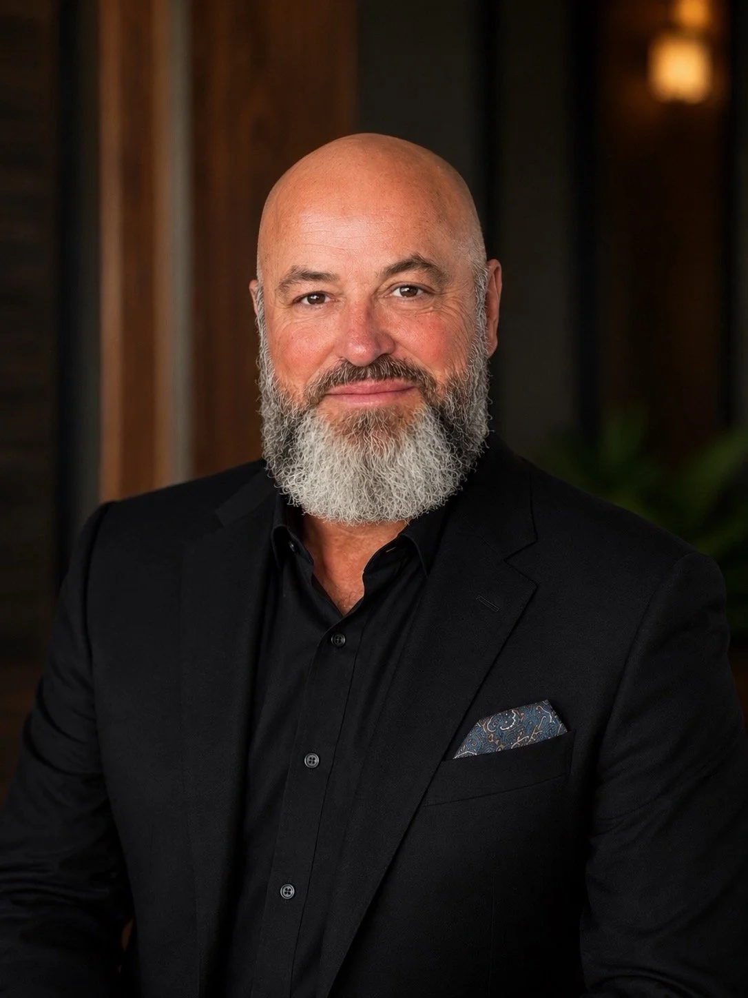 A bald man with a full gray beard, wearing a black suit and black shirt, posing indoors with warm lighting and dark background.