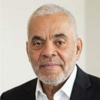 Close-up portrait of an older African American man with short gray hair and beard, wearing a black suit jacket over a white shirt, against a plain background.
