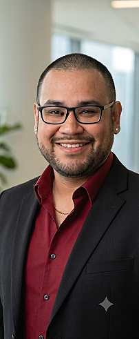 Smiling man wearing glasses, a black blazer, and a maroon shirt in an office setting.