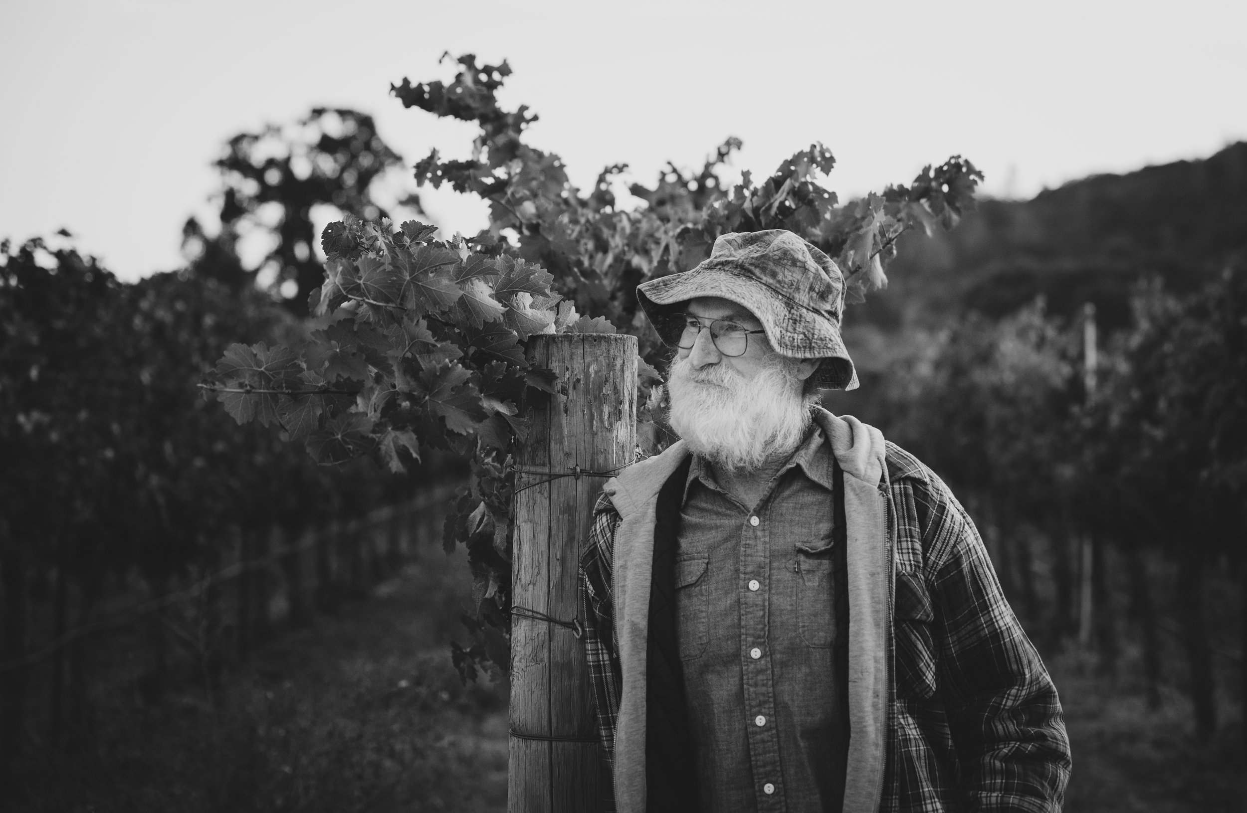 A black and white photo of an elderly man with a beard and glasses, wearing a bucket hat and checkered shirt, standing outdoors beside a wooden post with vines growing around it.