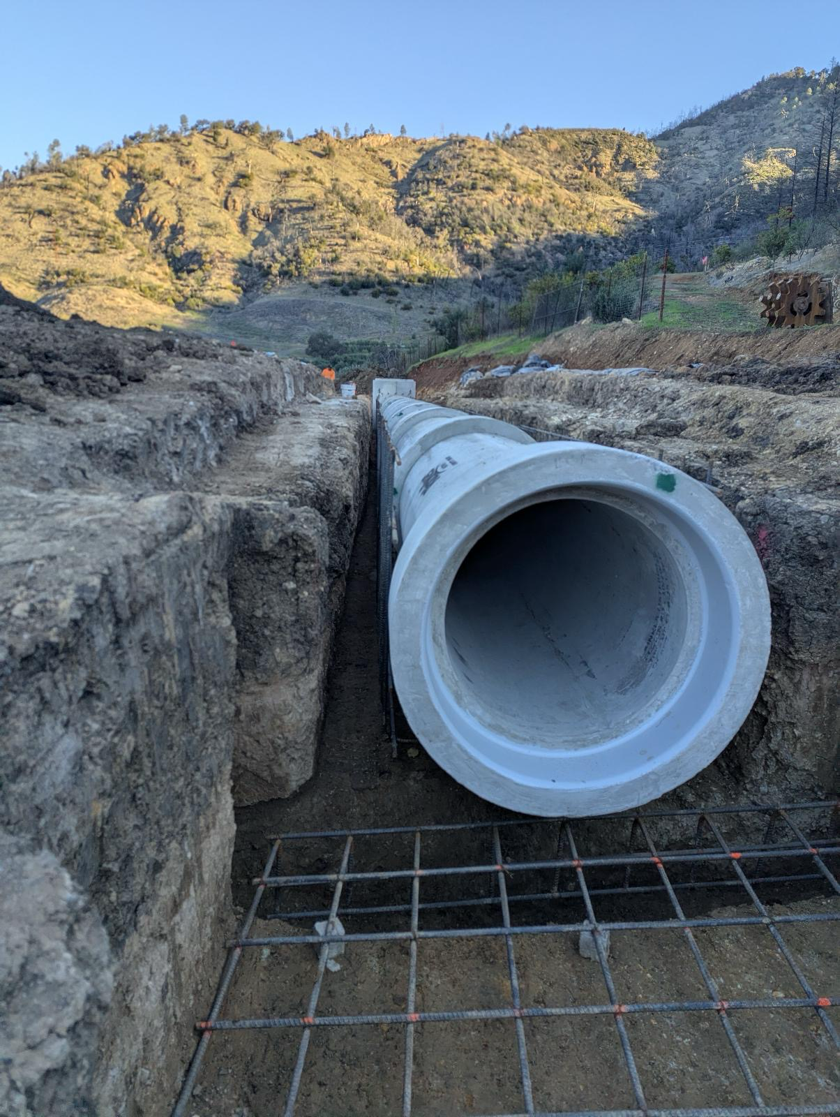 Construction site with a large pipe being installed in a trench surrounded by rocky soil, hills in the background, and a clear blue sky.