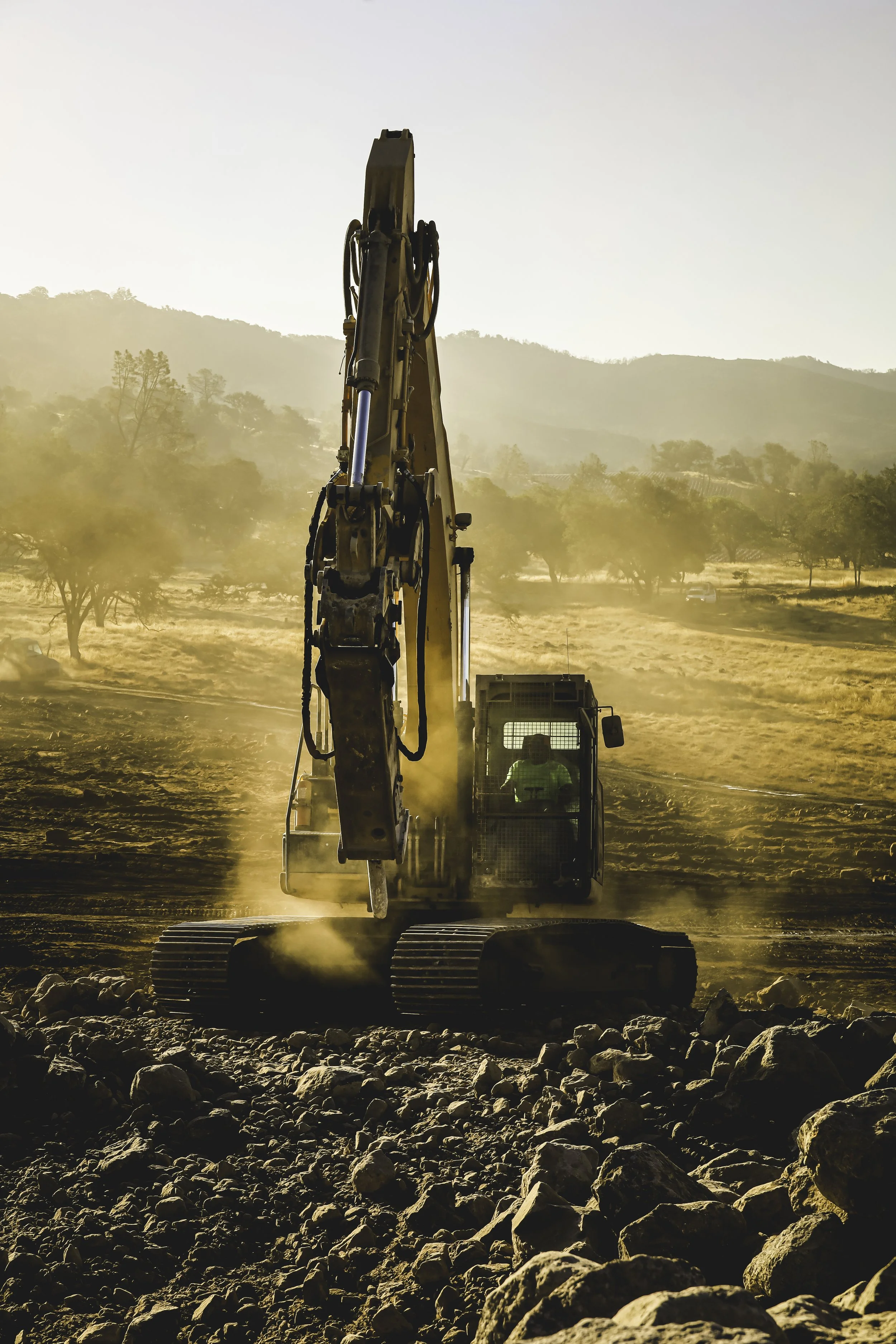 A large excavator operating on a construction site with rocky terrain, dust rising around it, and a backdrop of hills and trees during sunset or sunrise.