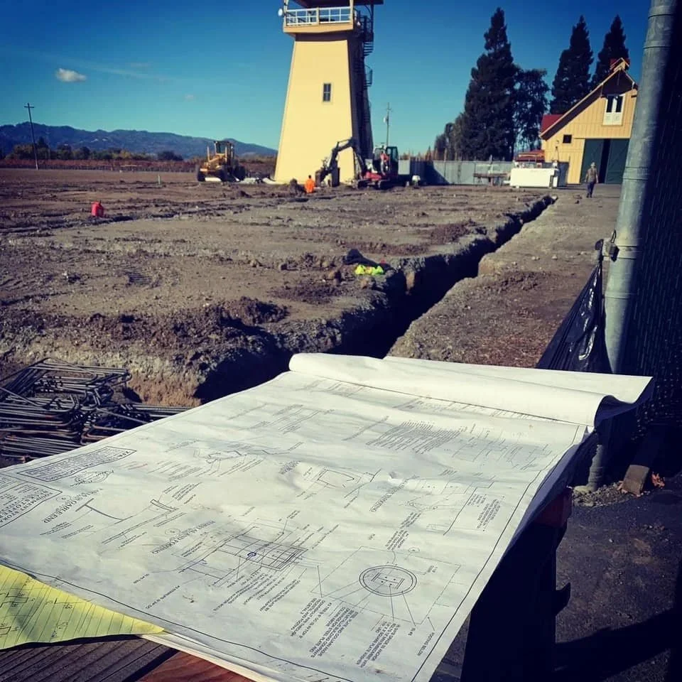 Construction site with blueprint plans on a table, excavation in progress, tower structure in the background, and a yellow house nearby.