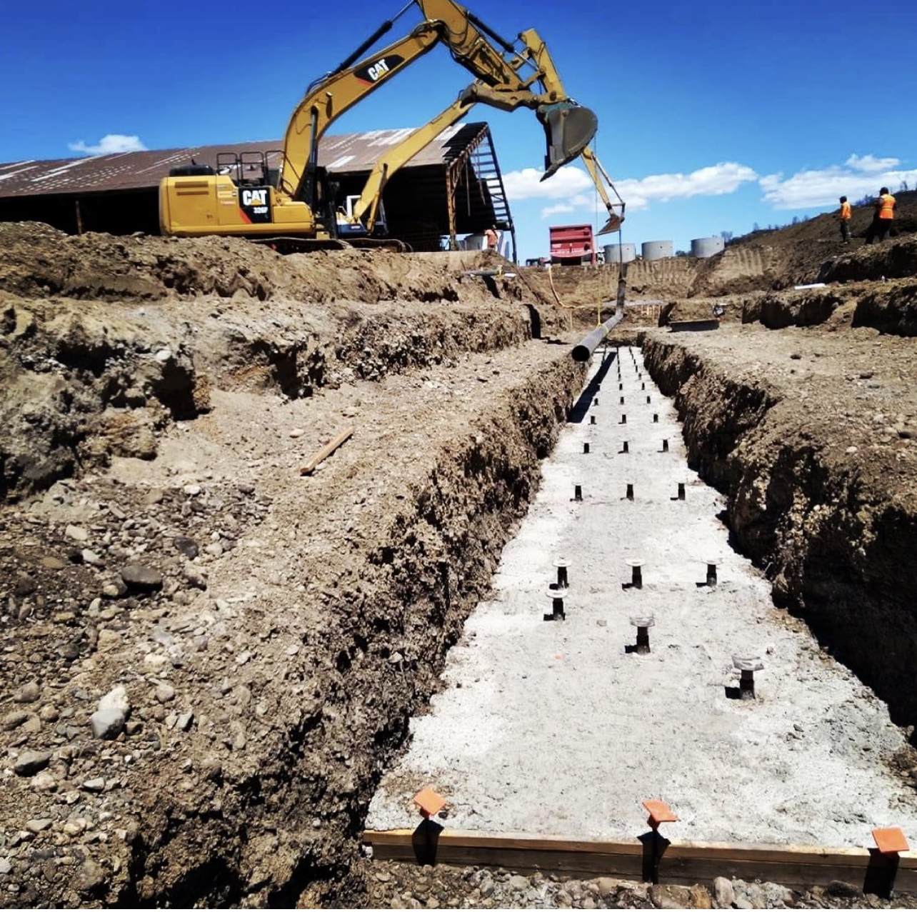 Construction site with a yellow excavator placing pipes in a trench. Several workers are visible in safety vests, working in the background under a clear blue sky.