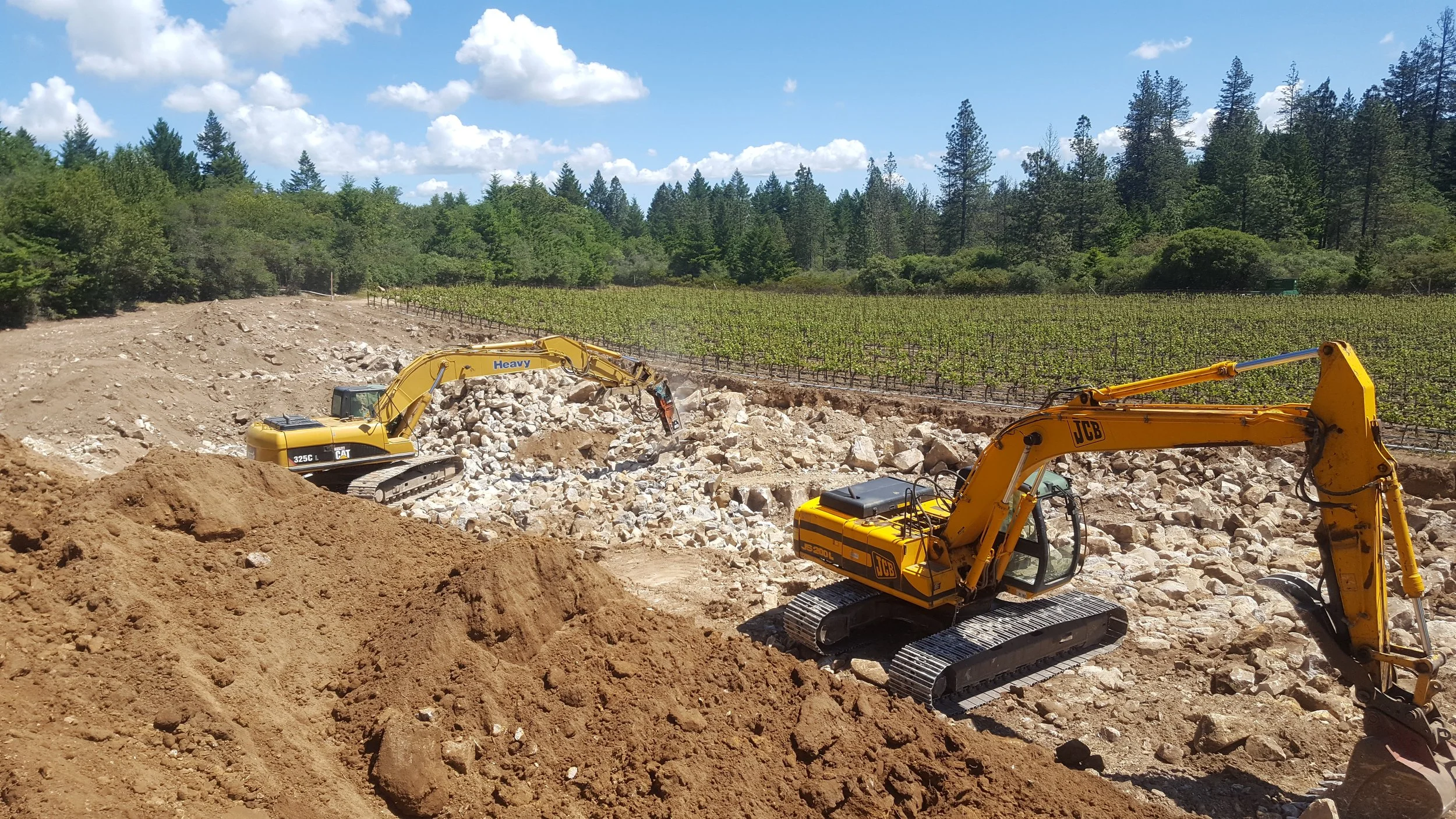Construction site with two yellow excavators working on rocky terrain, with a vineyard and forest in the background under a partly cloudy sky.