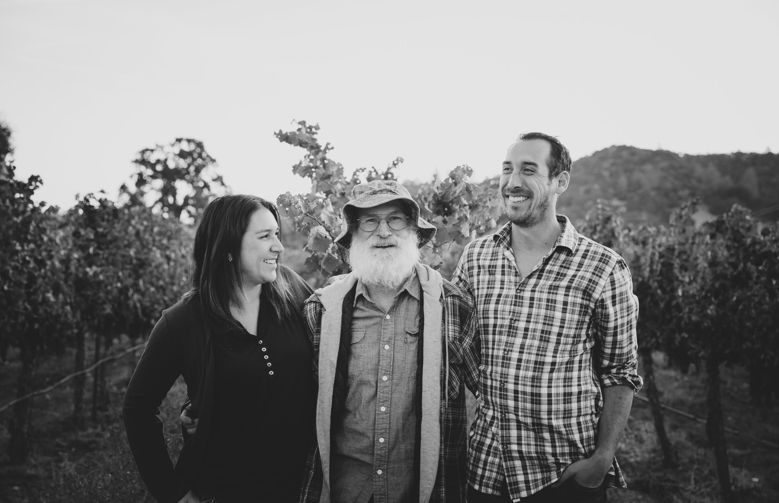 A group of four people standing outdoors, smiling, with trees and hills in the background, in black and white.