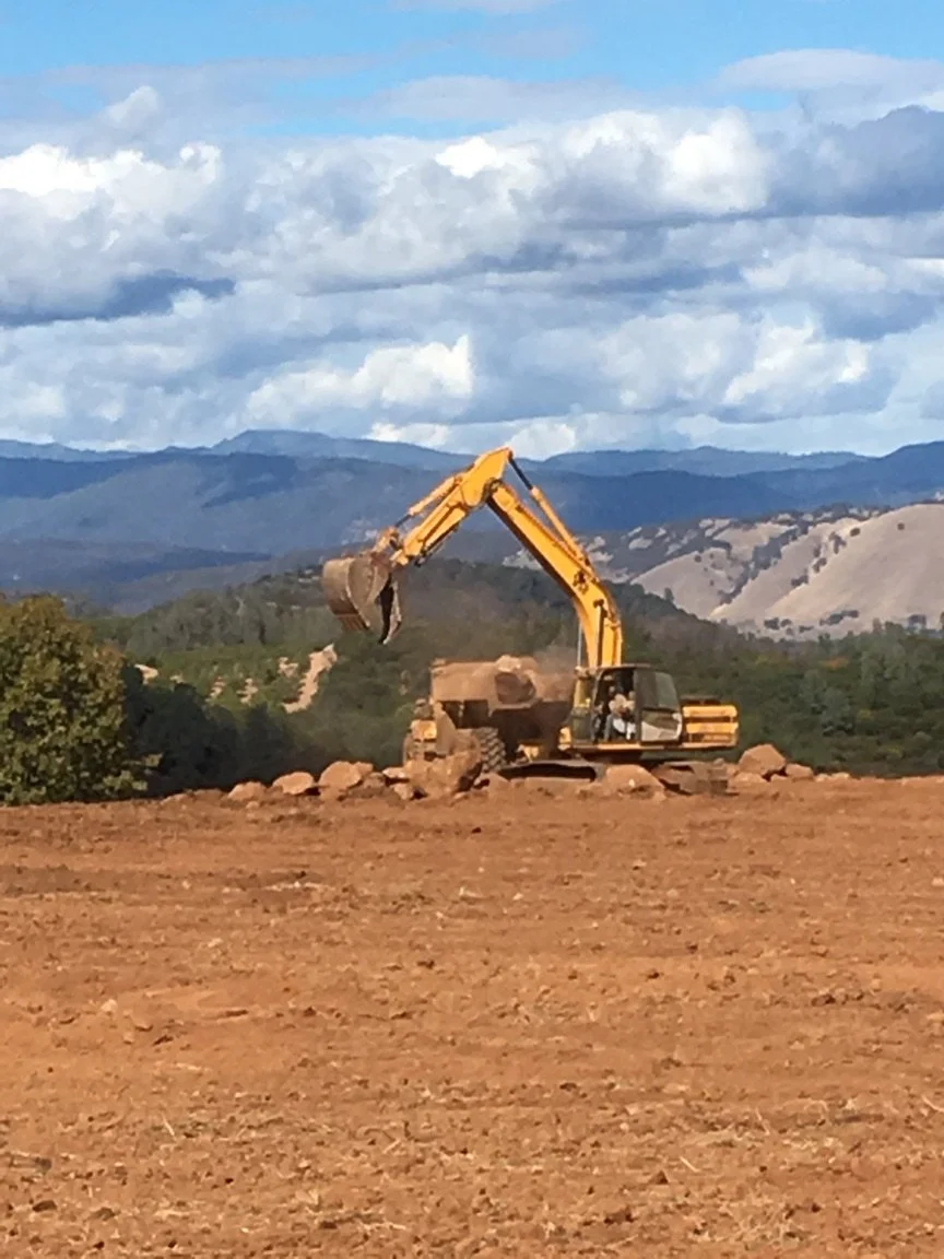 A yellow excavator working on a construction site with mountains and cloudy sky in the background.