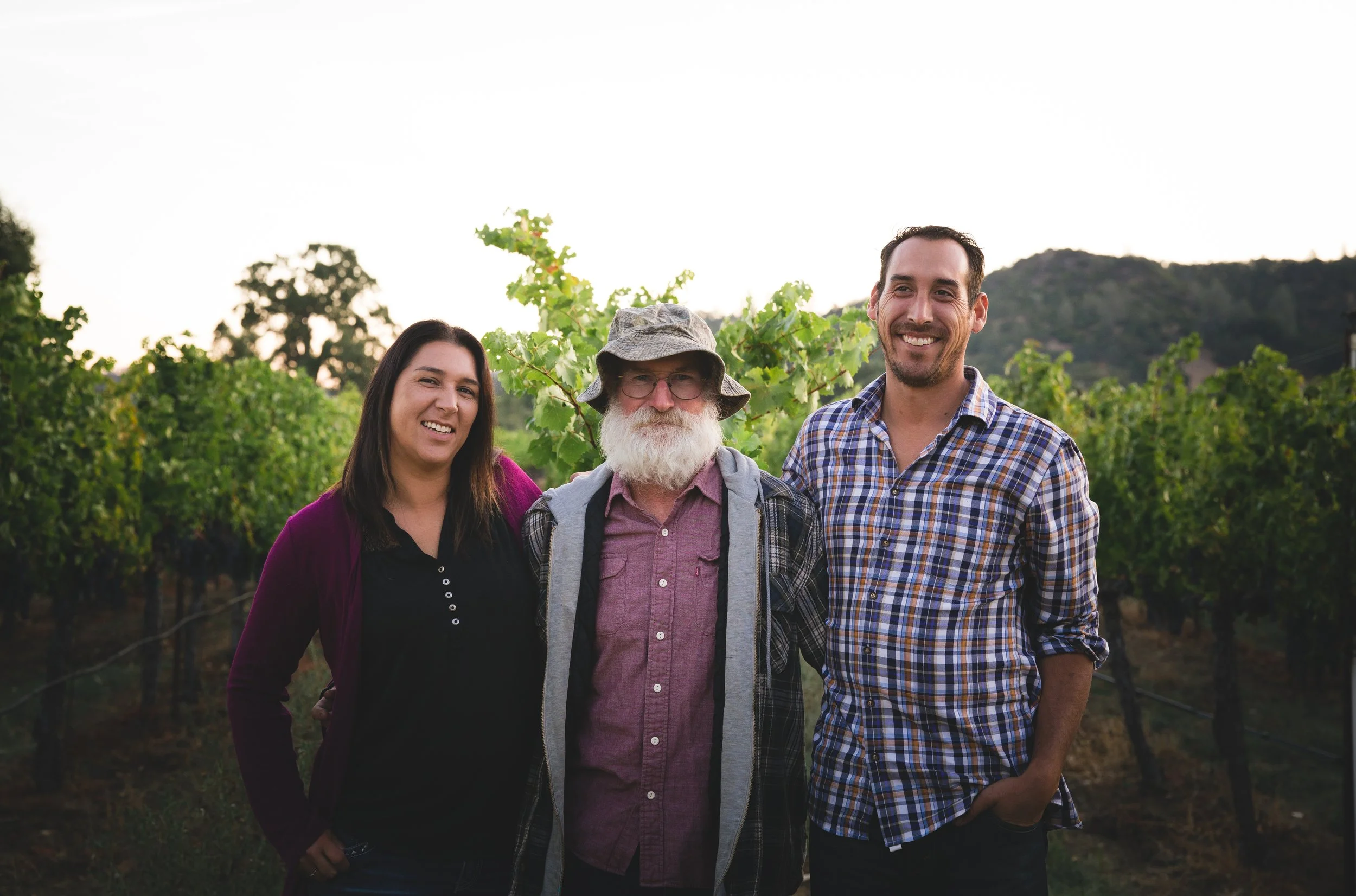Four people standing outdoors in a vineyard during daylight, smiling at the camera. The group includes a woman with long dark hair, an older man with a white beard and glasses wearing a hat, and two men with short dark hair, one wearing a plaid shirt.