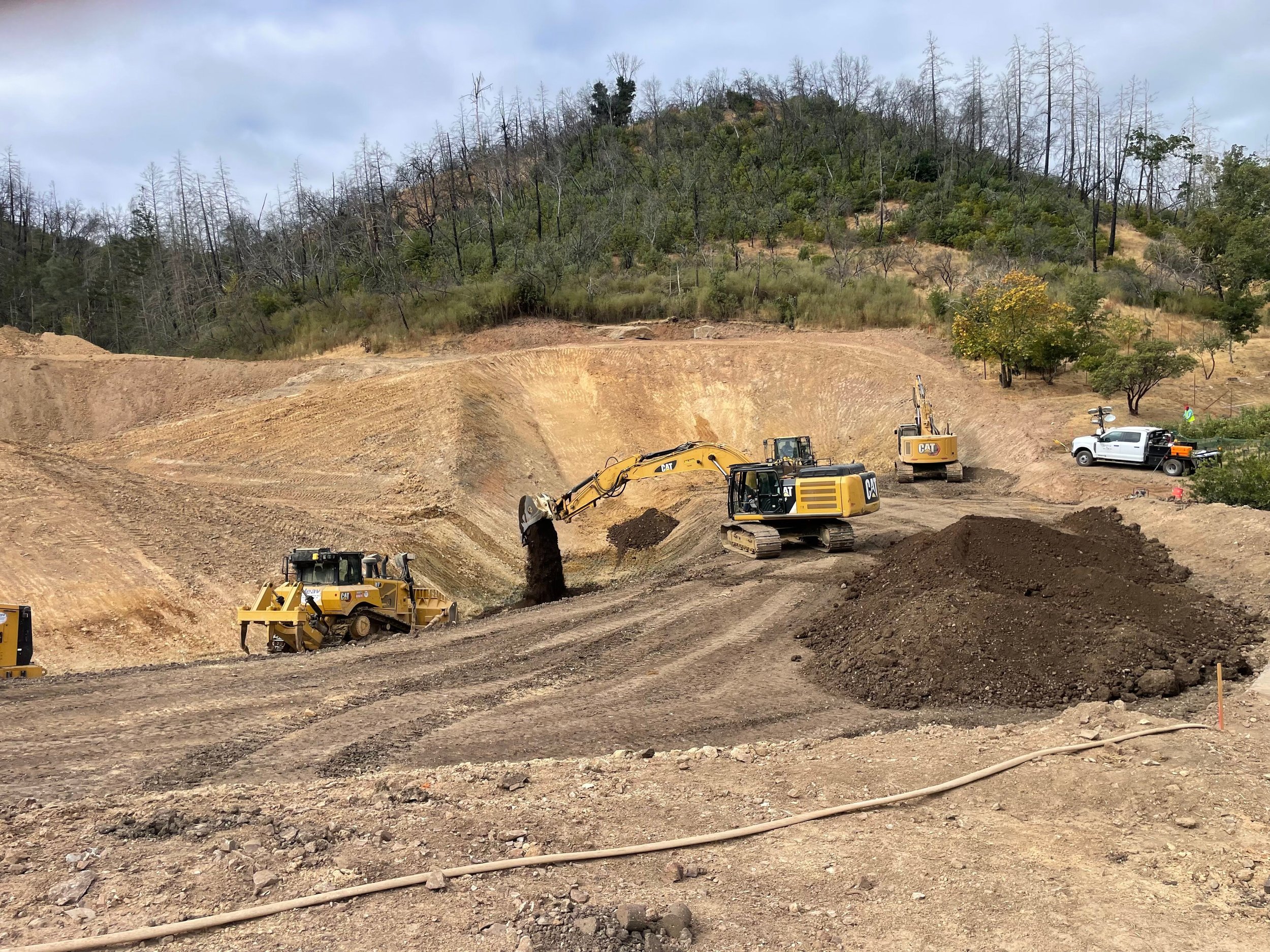 Construction site on a hillside with excavators, a white utility truck, and dirt piles, with burnt trees and green vegetation on the hill in the background.