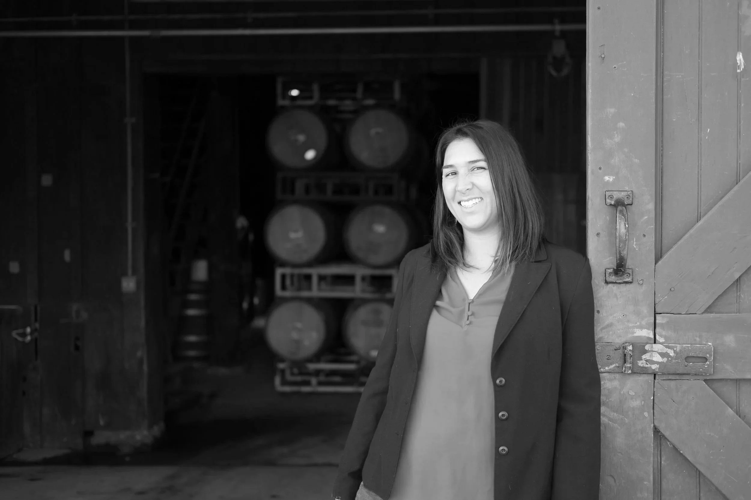 Black and white photo of a smiling woman with long dark hair, wearing a blazer, standing partly inside a wooden building with barrels in the background.
