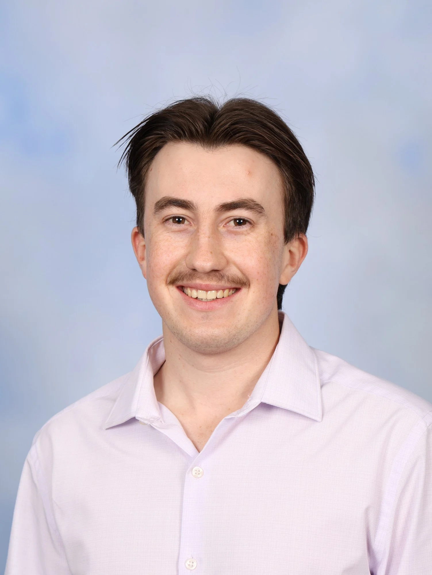 Portrait of a young man with brown hair, light skin with freckles, smiling, wearing a light pink button-up shirt, against a light blue background.