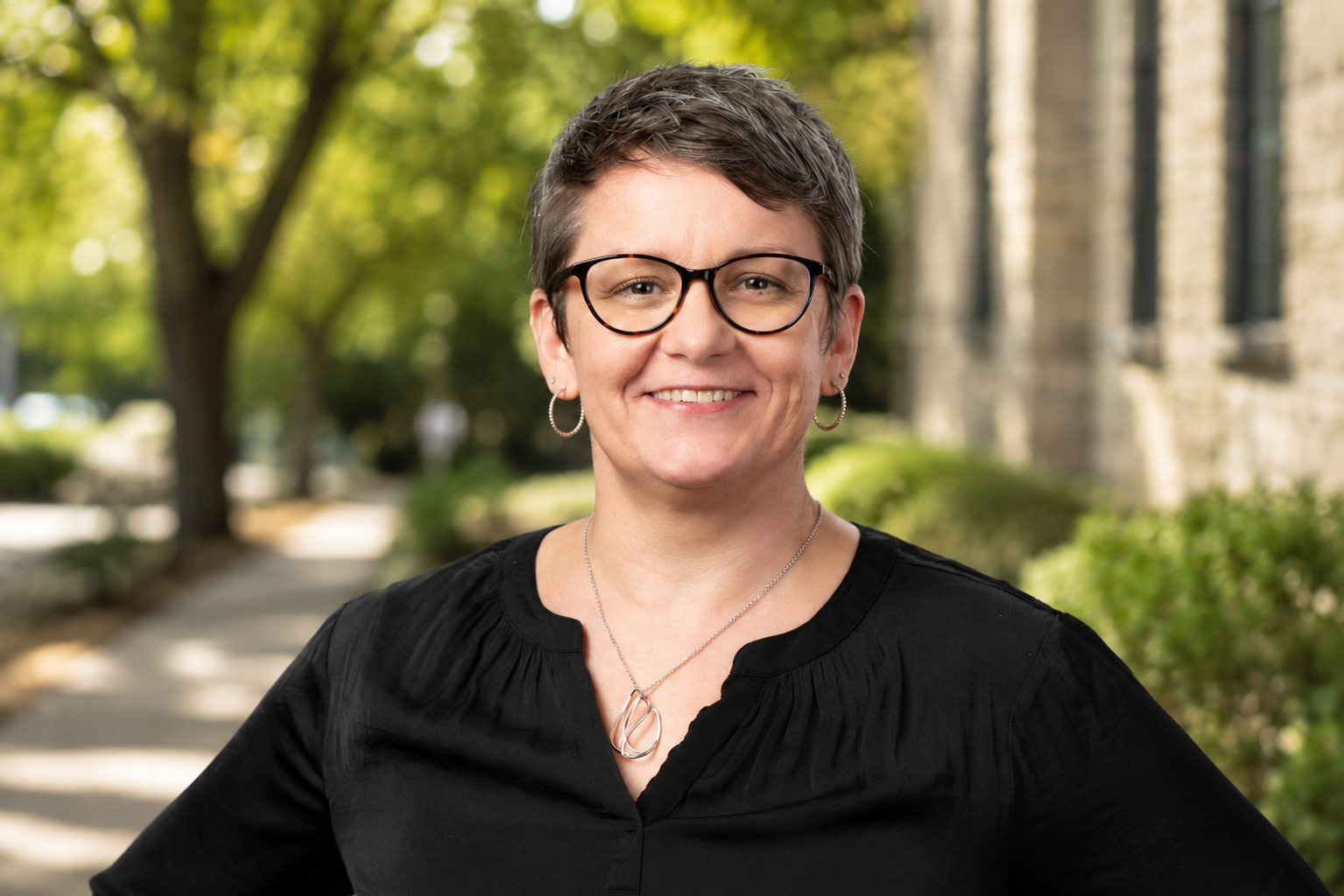 A smiling woman with short gray hair and glasses, wearing a black top, standing outdoors on a sidewalk with trees and a stone building in the background.