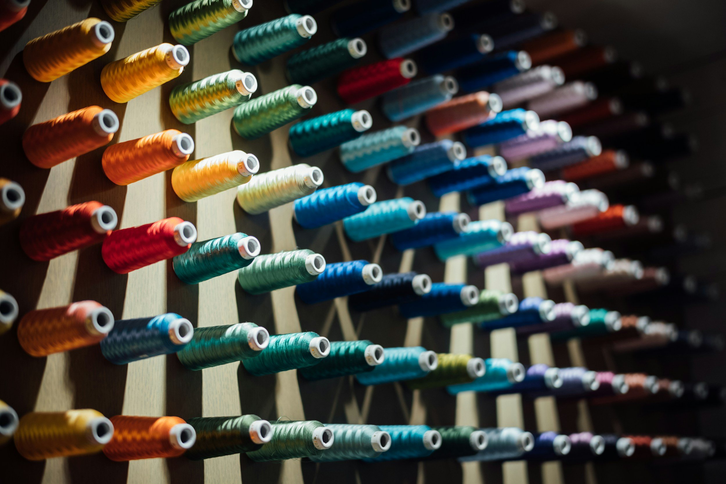 Close-up of an organized display of colorful sewing thread spools on a wooden rack, featuring various shades including yellow, orange, green, blue, purple, and red.
