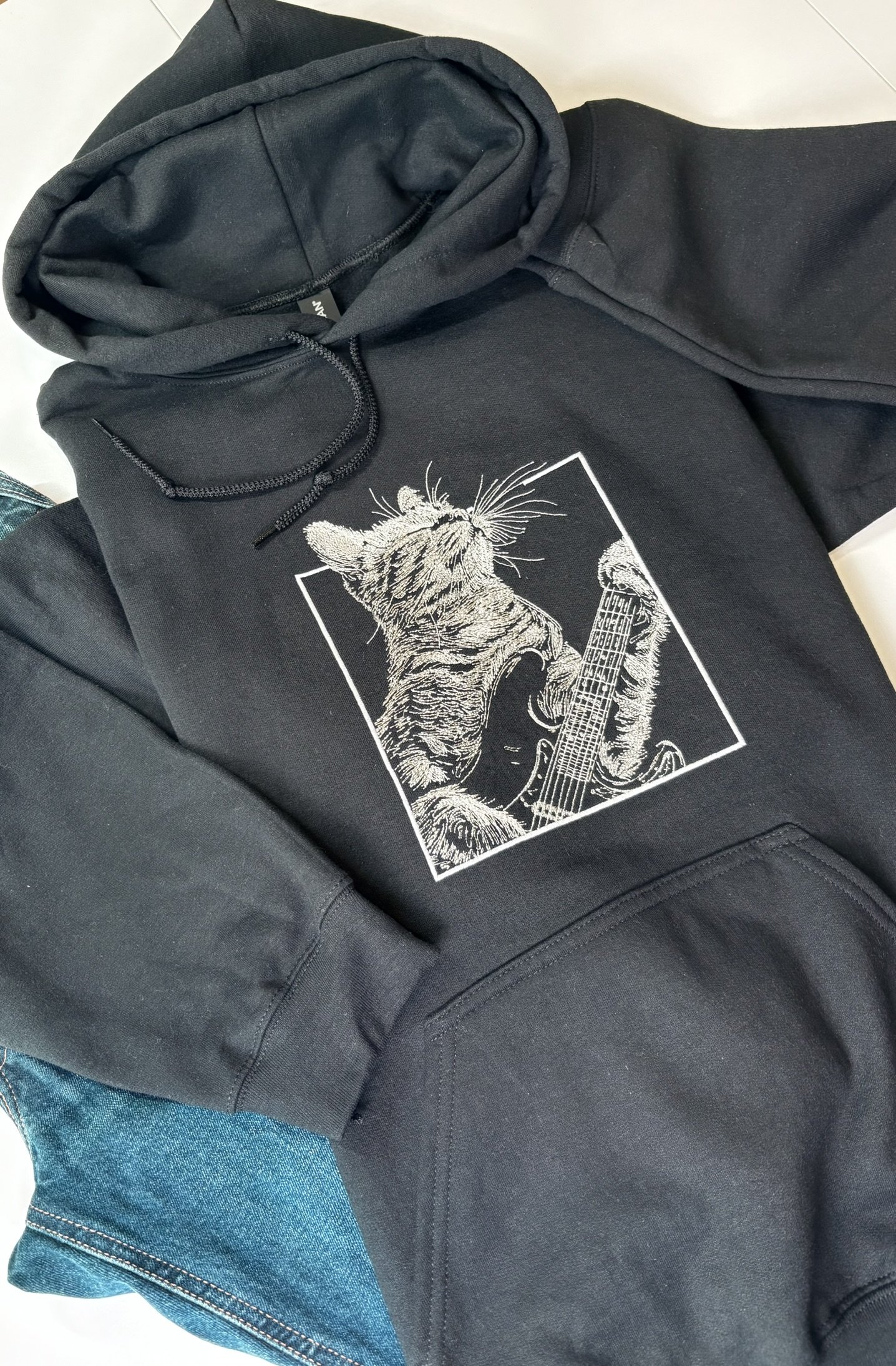 A black hoodie with a gray and white embroidered cat holding a guitar on the front