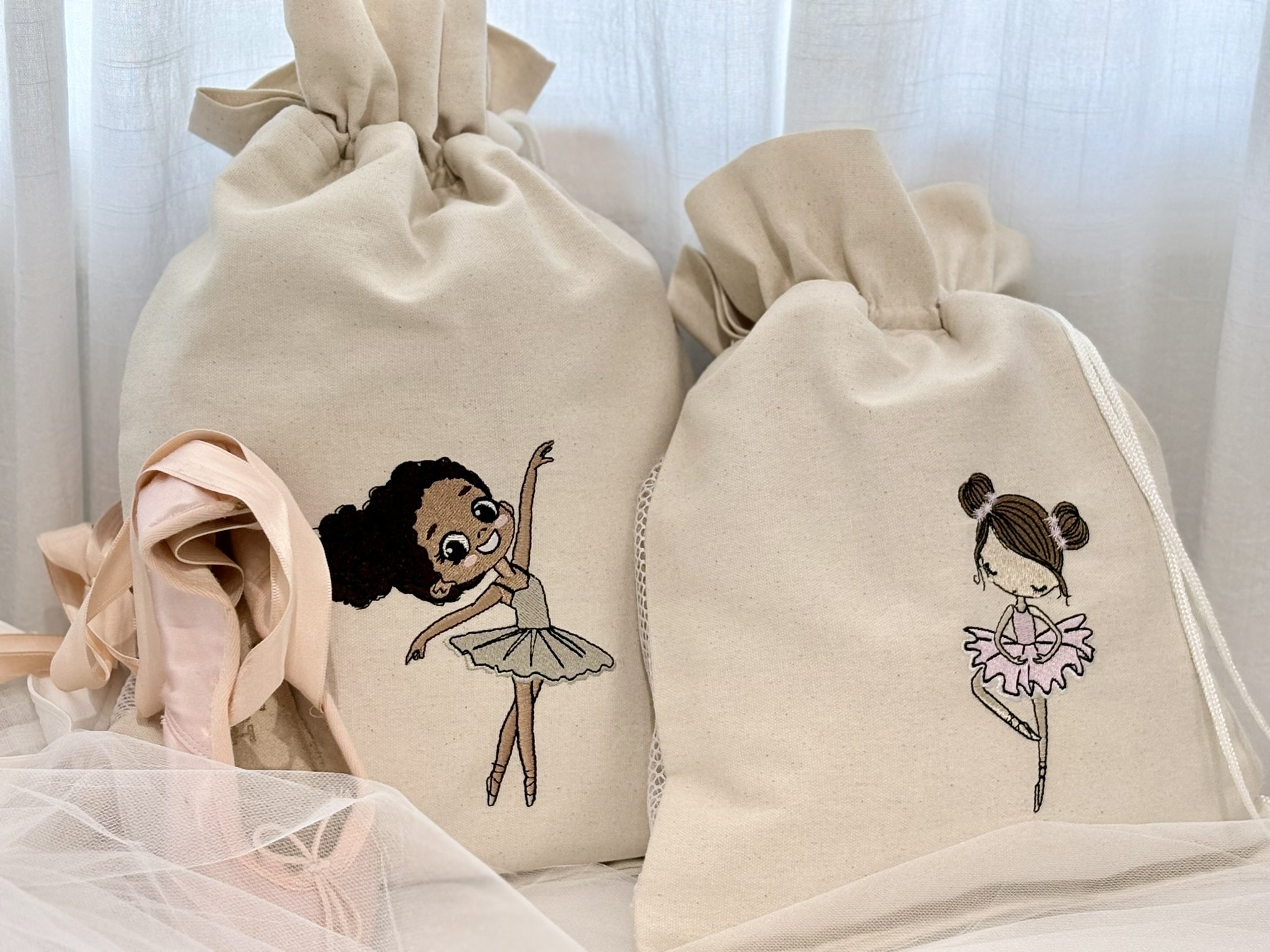 Close-up of beige canvas ballet shoe bags with embroidered ballet dancers, one with curly hair and the other with pigtails, hanging against a sheer curtain background.