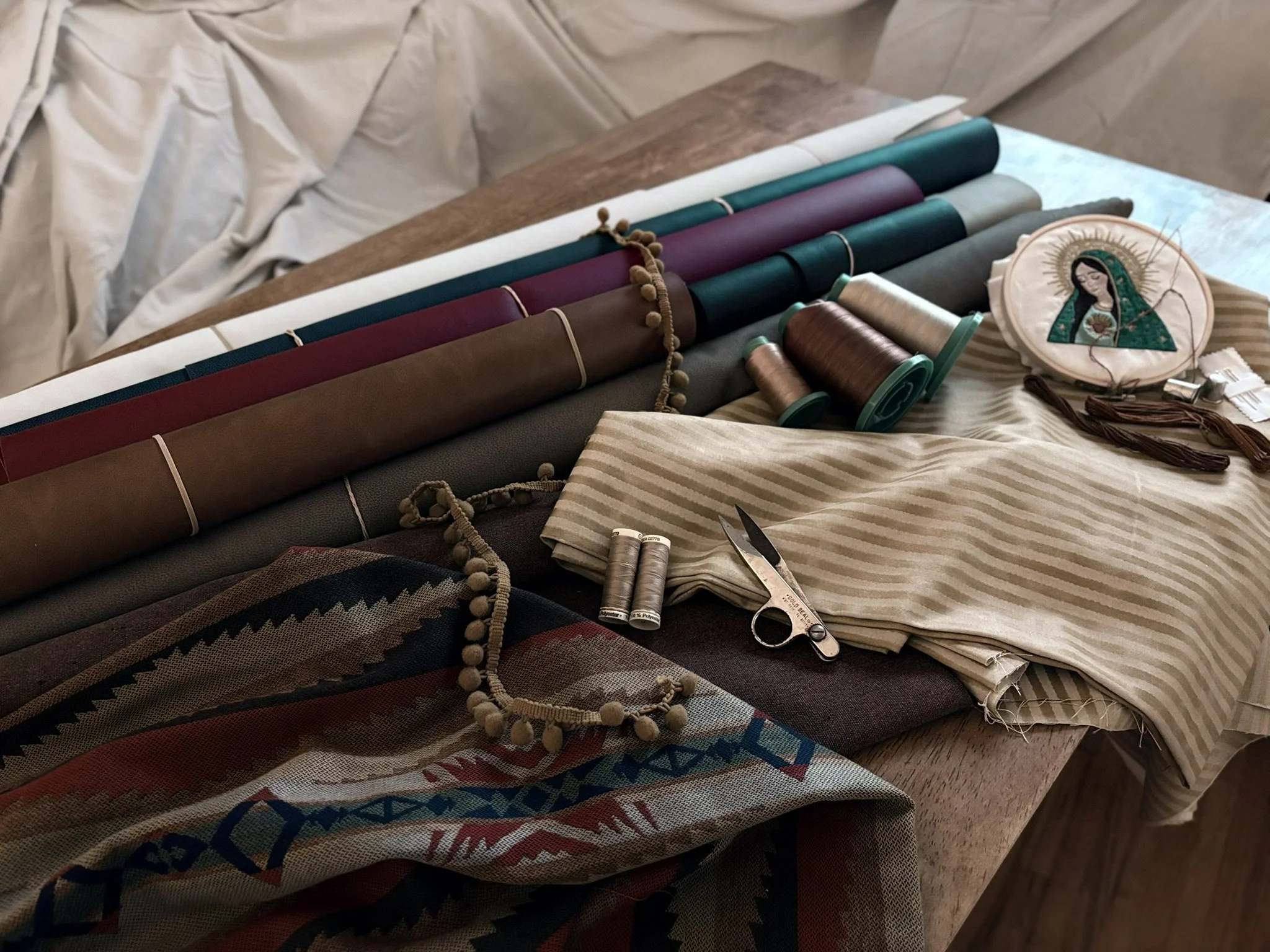 A table covered with various fabrics, including patterned, striped, and solid colors, along with spools of thread, a pair of scissors, and decorative beads, all in a craft or sewing setting.