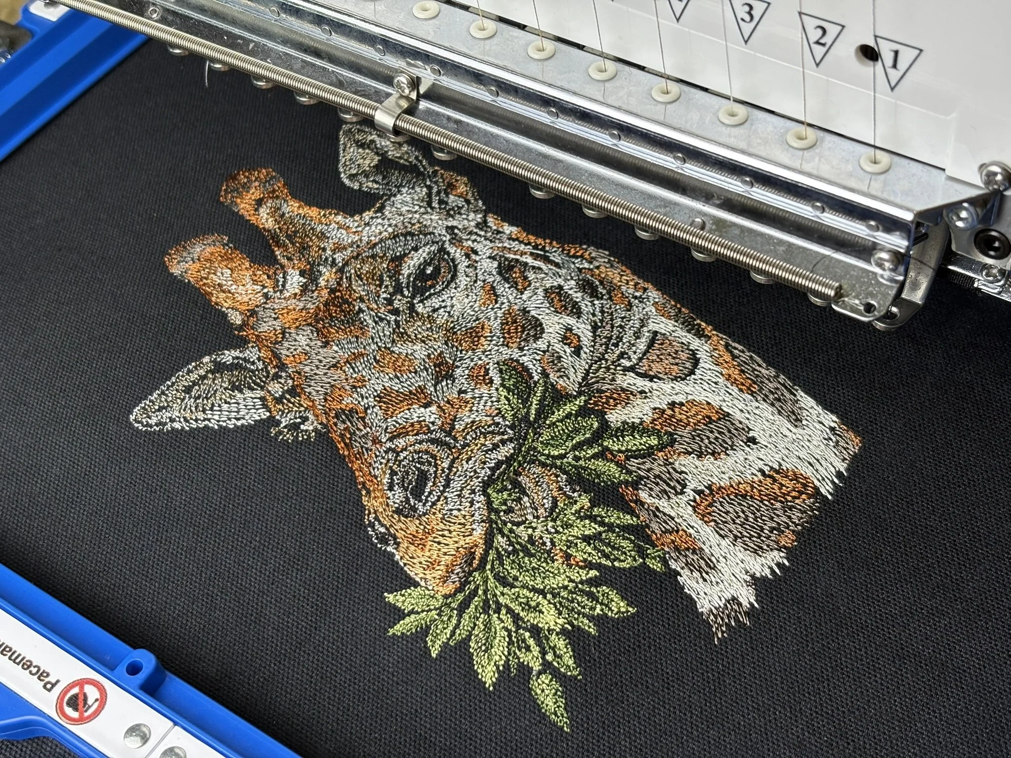 Close-up of an embroidery machine stitching a detailed design of a giraffe's face with orange, black, white, and green threads on a black fabric background.