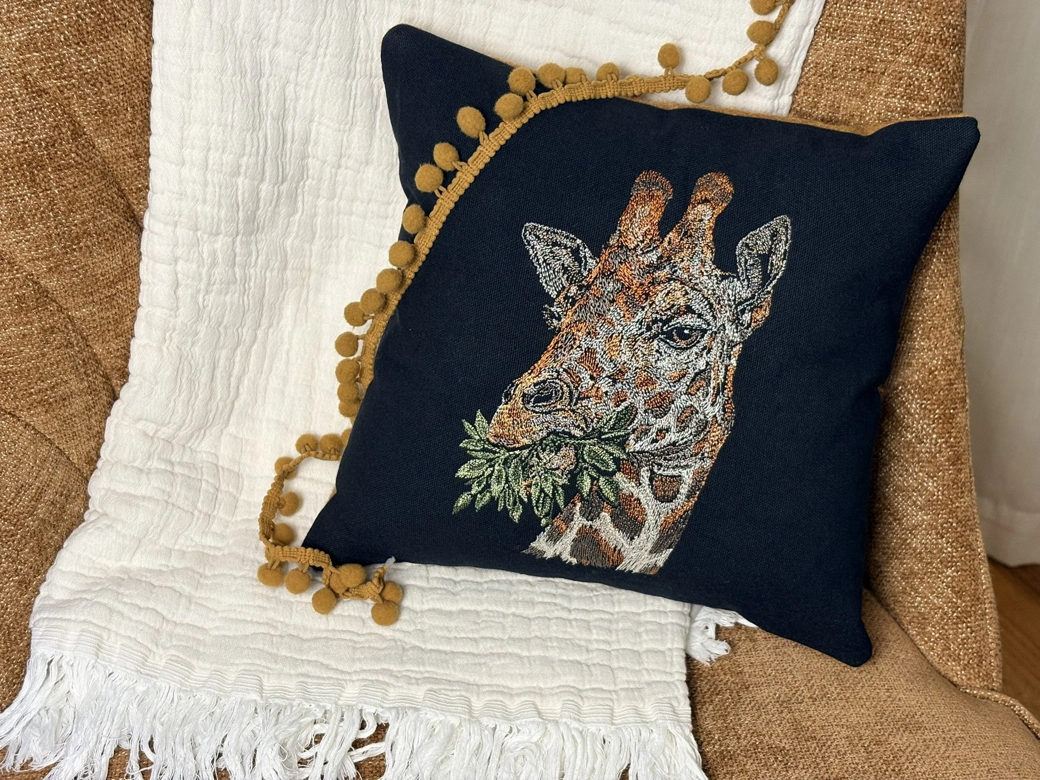 A decorative pillow with a giraffe's face embroidered on dark fabric, outlined with tapestry stitches, and trimmed with a yellow pom-pom fringe, placed on a white textured throw blanket on a brown couch.