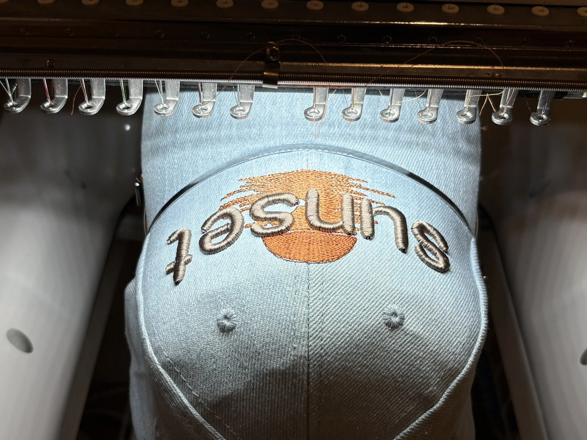 Close-up view of embroidery machine stitching a fabric with the text 'Sunset' in a puff lettering dsign and an embroidered orange sun or sunset design.
