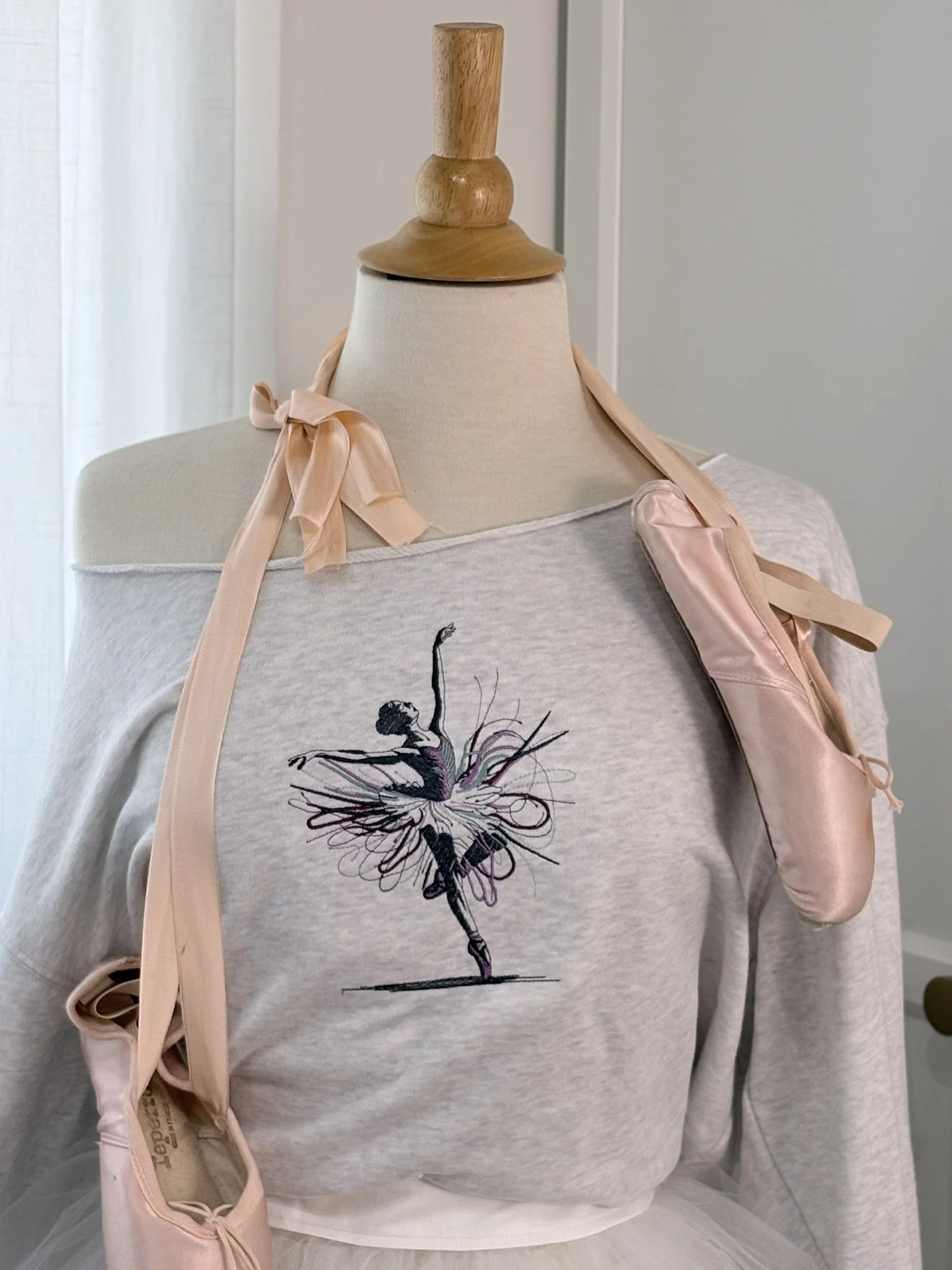 A mannequin displays a ballet dancer embroidered on a light gray sweatshirt, with a ballet dancer illustration and a ballet pose in black and purple thread. The mannequin has a pink satin ballet shoe draped over its shoulder and a wooden top handle.