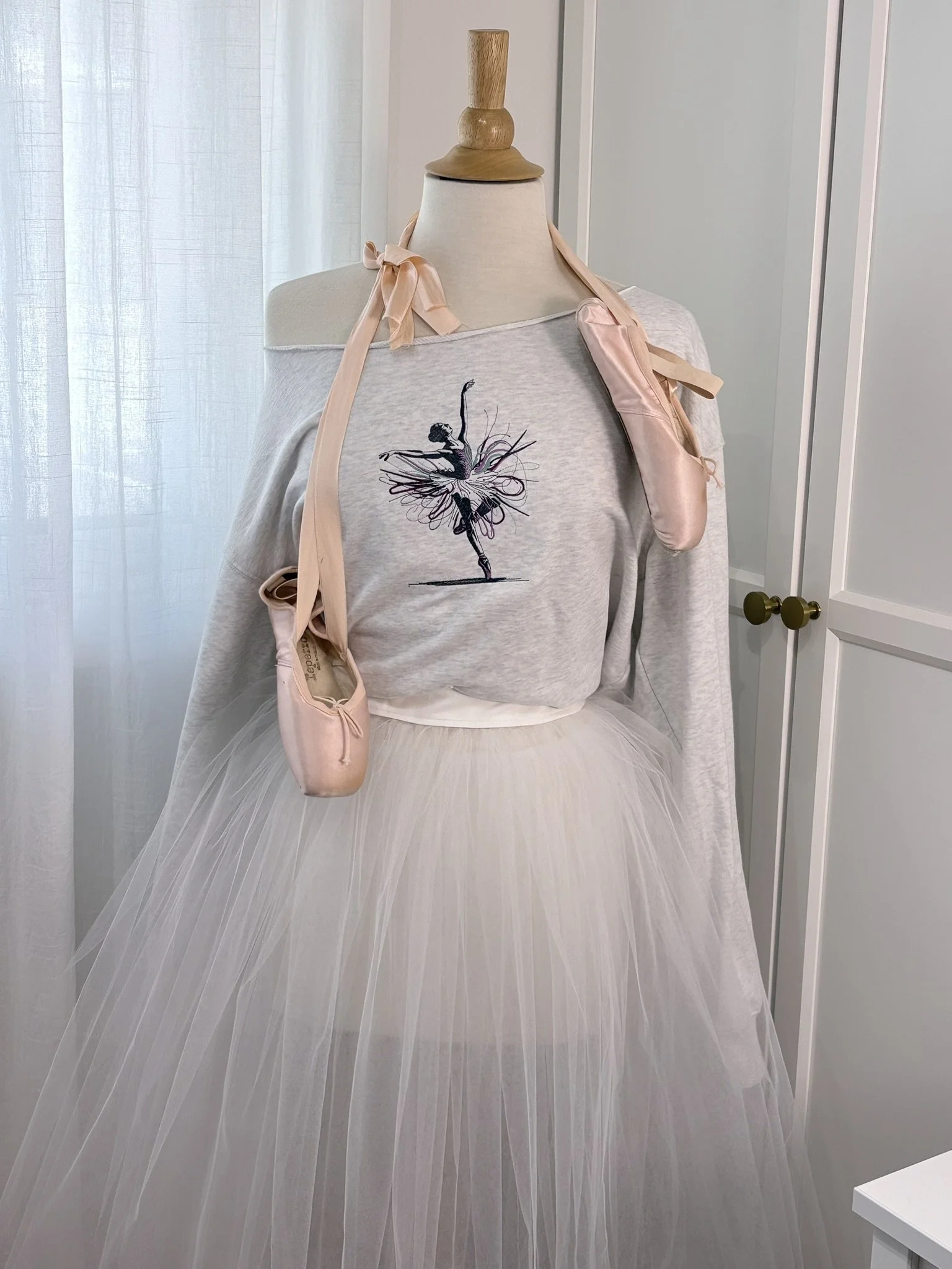 A mannequin dressed in a ballet-themed outfit, with a light grey sweatshirt featuring an embroidered black ballerina silhouette, a white tutu, pink ballet slippers draped over shoulders of the mannequin stand