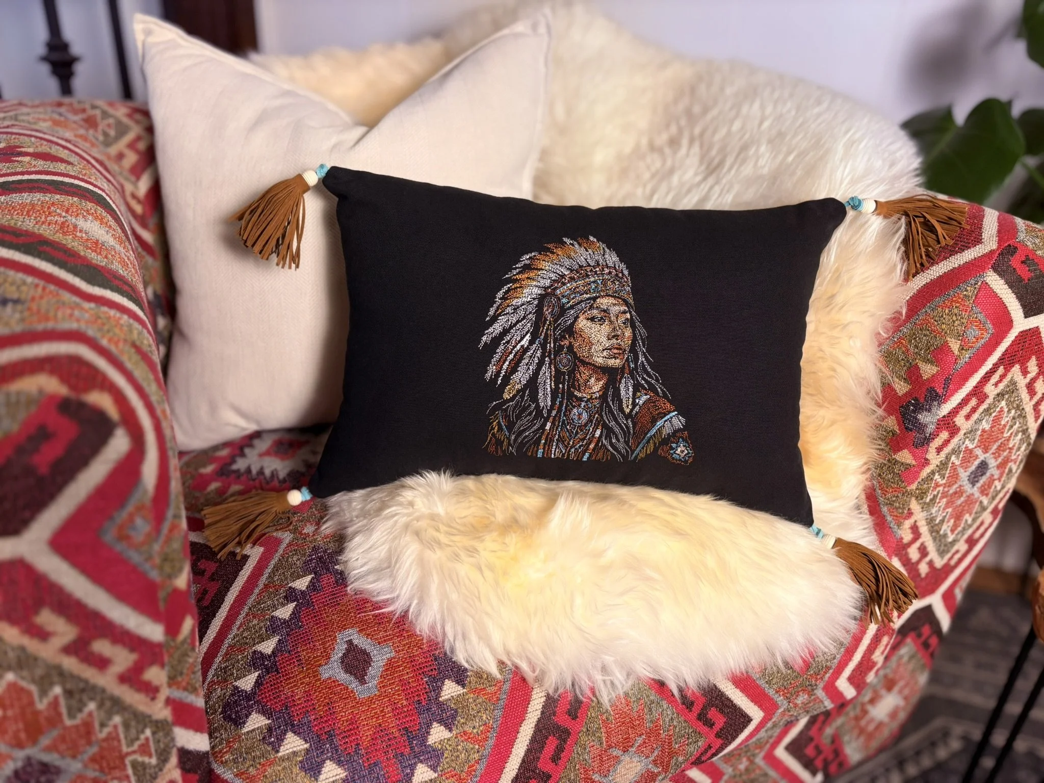 A decorative black embroidered pillow with a Native American chief in headdress, resting on a fluffy white cushioned surface, surrounded by patterned cushions with fringes and tassels.