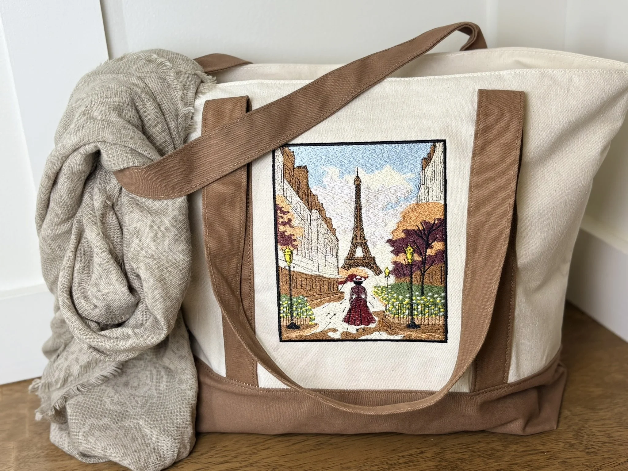 A canvas tote bag with a zipper, featuring an embroidered scene of Paris including the Eiffel Tower, with a beige and brown color scheme. A beige scarf is draped over the bag's handle.