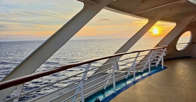 On the deck of a cruise ship at sunset, with a view of the ocean and the horizon.