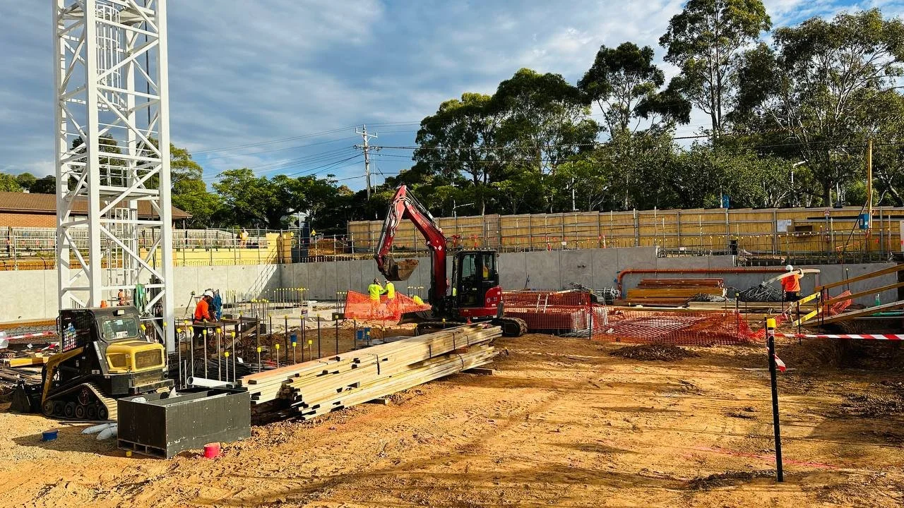 Construction site with workers, excavator, materials, and equipment under a cloudy sky.