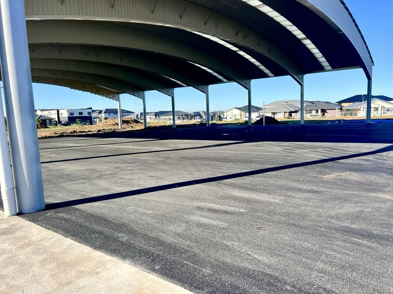 Empty parking lot with a large covered structure and residential houses in the background under a clear blue sky.