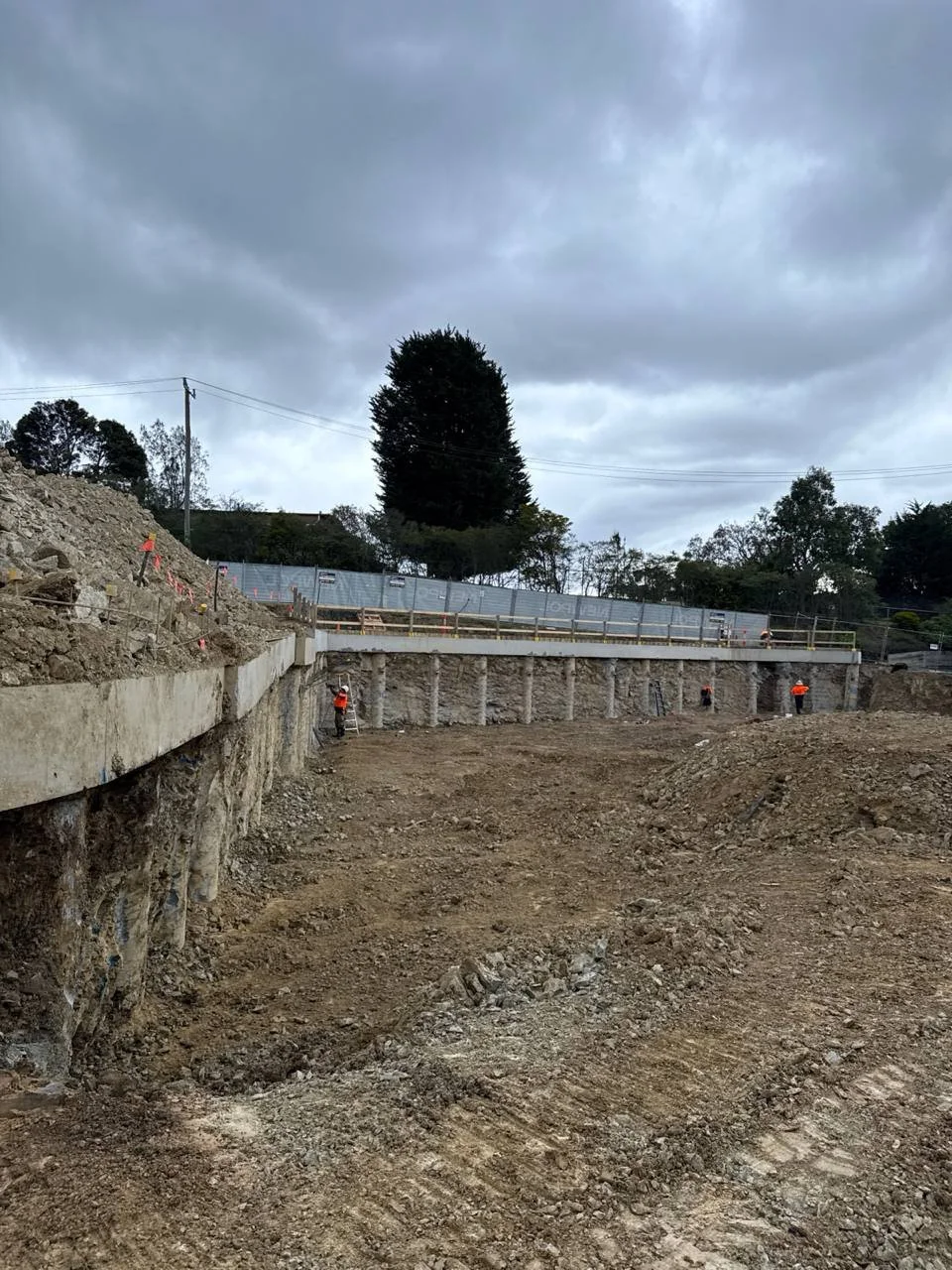 Construction site with workers in safety vests and helmets, dirt ground, and a partially built retaining wall under a cloudy sky.