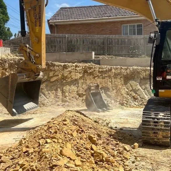 Construction site with a yellow excavator digging and a pile of dirt and rocks in the foreground. A house with a brick exterior and a wooden fence is visible in the background.