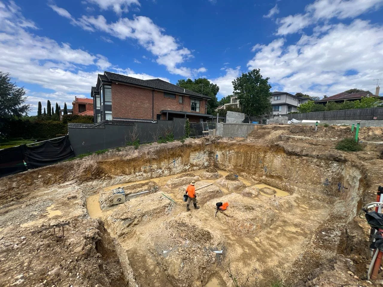 Construction site with an excavated foundation being prepared for building, worker wearing orange vest and hat working on the site, residential houses and a partly cloudy sky in the background.