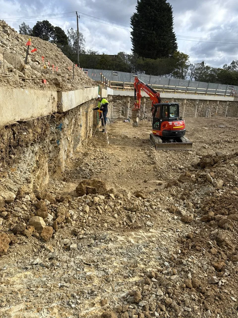 Construction site with excavator and workers during earthwork, with rocky soil, safety barriers, and trees in the background.
