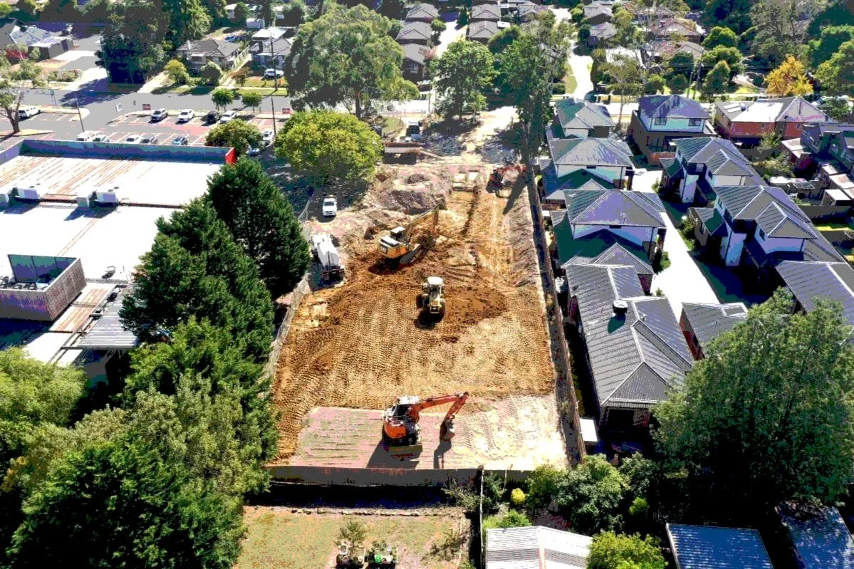 An aerial view of a construction site in a residential neighborhood with excavators working on cleared land, surrounded by houses and trees.