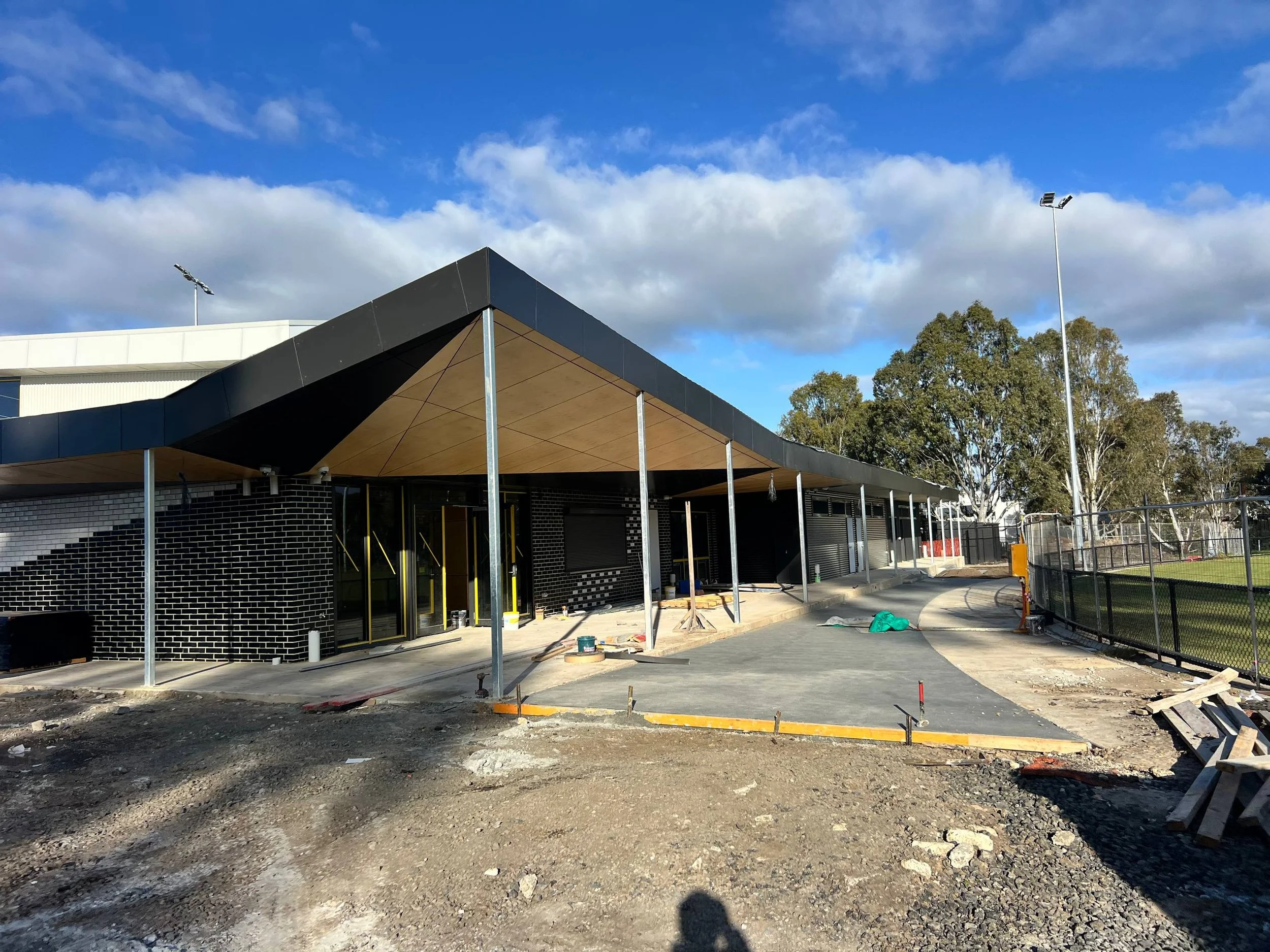 Building under construction with a black roof, brick walls, and construction materials around. Construction site with dirt ground and some orange safety barriers, trees and a blue sky with clouds in the background.