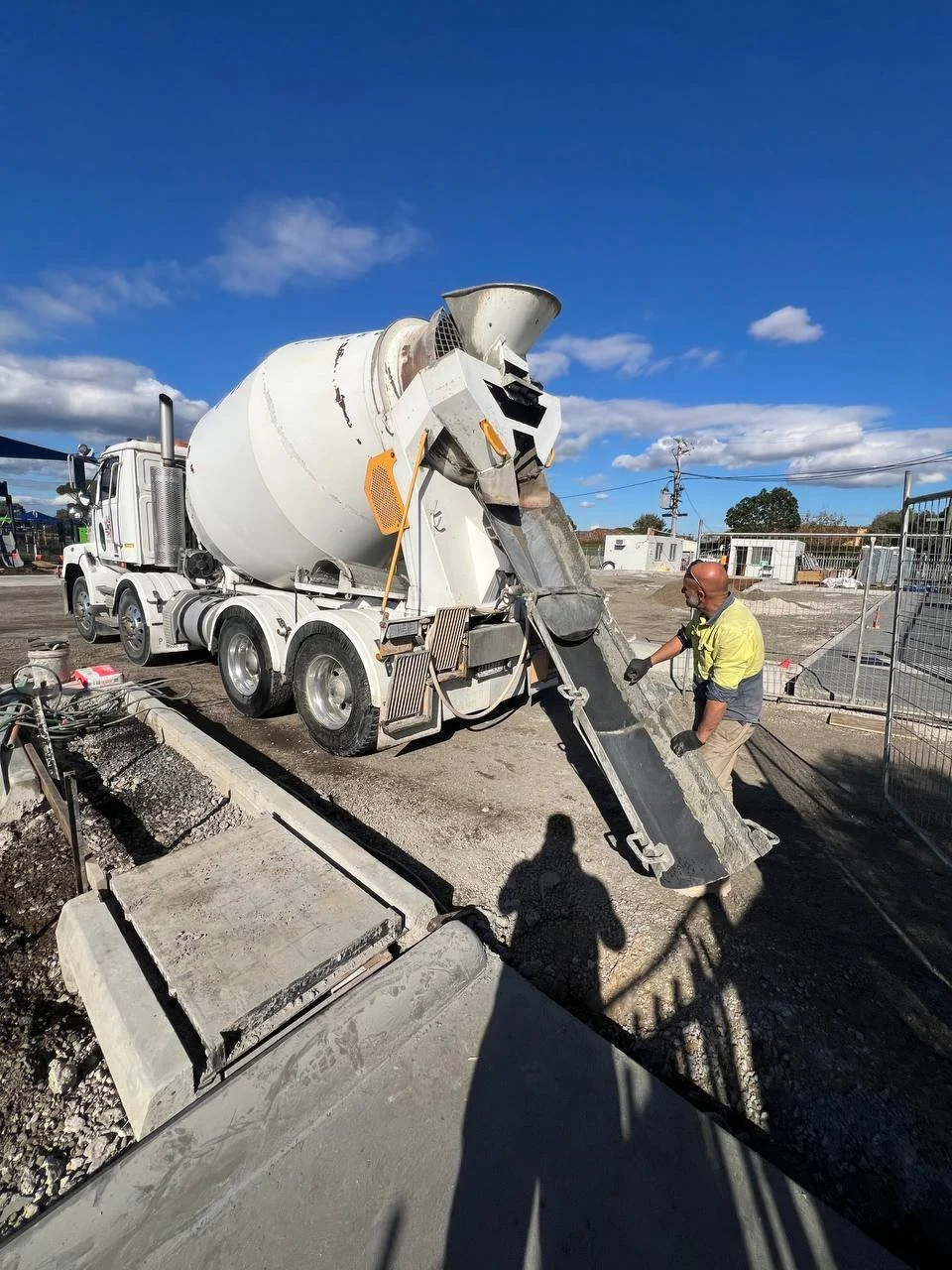 A construction worker operating a concrete mixer truck pouring concrete on a construction site with a blue sky and some clouds overhead.
