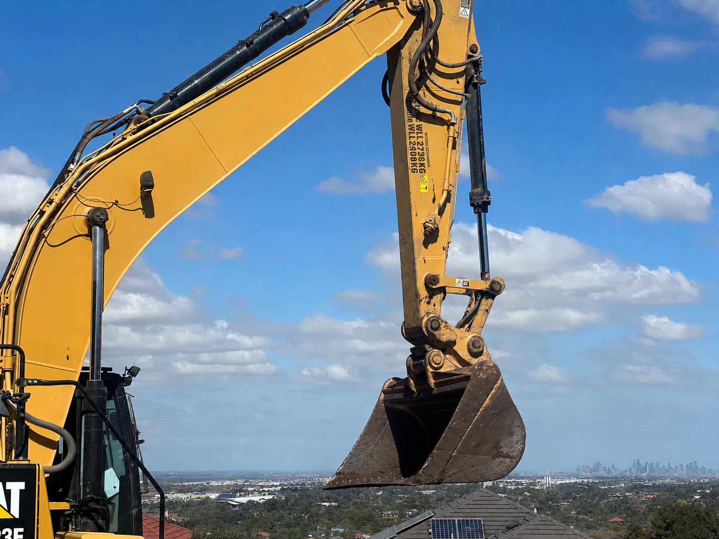 A yellow construction excavator with a bucket attachment working in an area with a clear blue sky and some clouds, with a city skyline visible in the background.