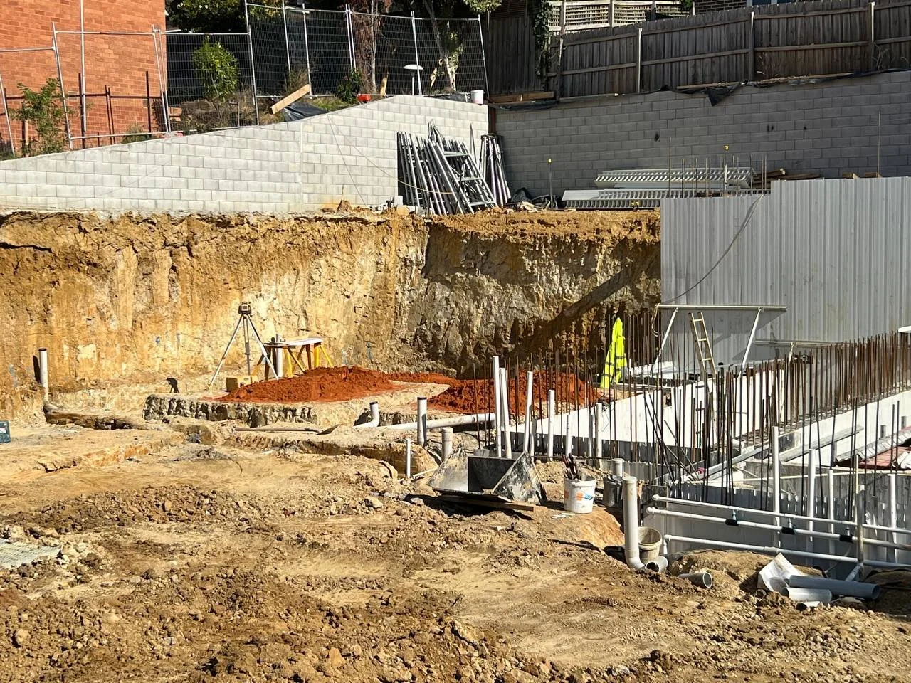 Construction site with dug earth, PVC pipes, metal scaffolding, a high-visibility safety vest hung on a ladder, and construction tools and materials scattered around.