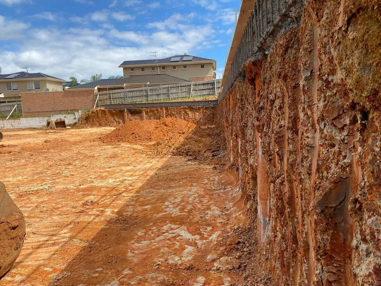 Construction site with exposed dirt and foundation, with houses and a fence in the background under a partly cloudy sky.