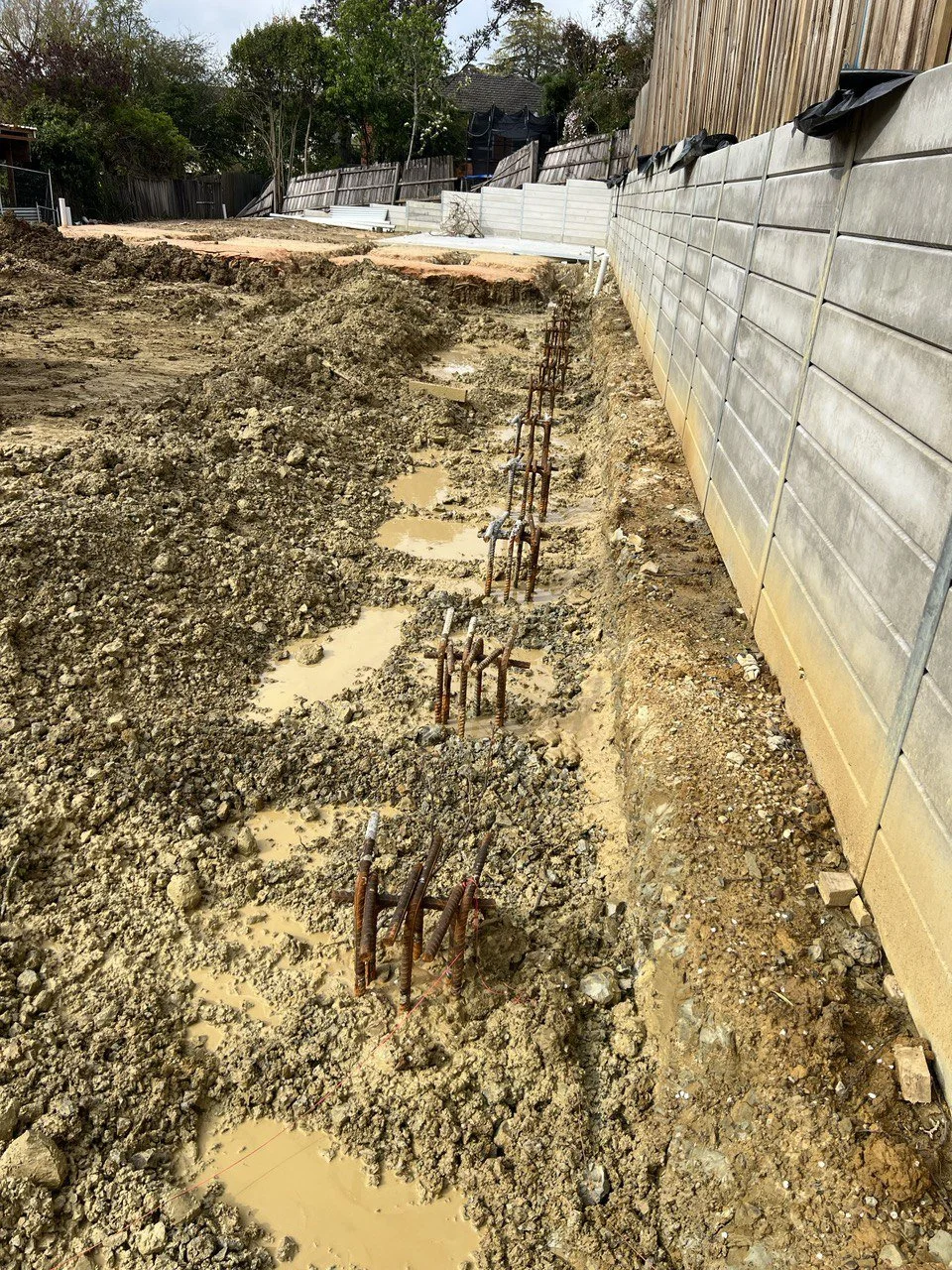 Construction site with exposed rebar and muddy ground, alongside a concrete fence.
