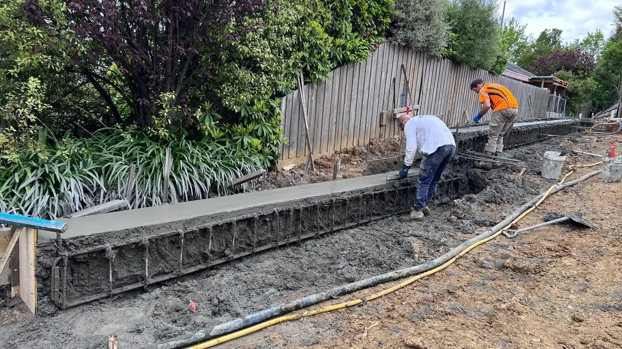 Two construction workers are building a concrete wall along a dirt path next to a wooden fence and greenery. One worker is smoothing the concrete, while the other is working further along the wall.